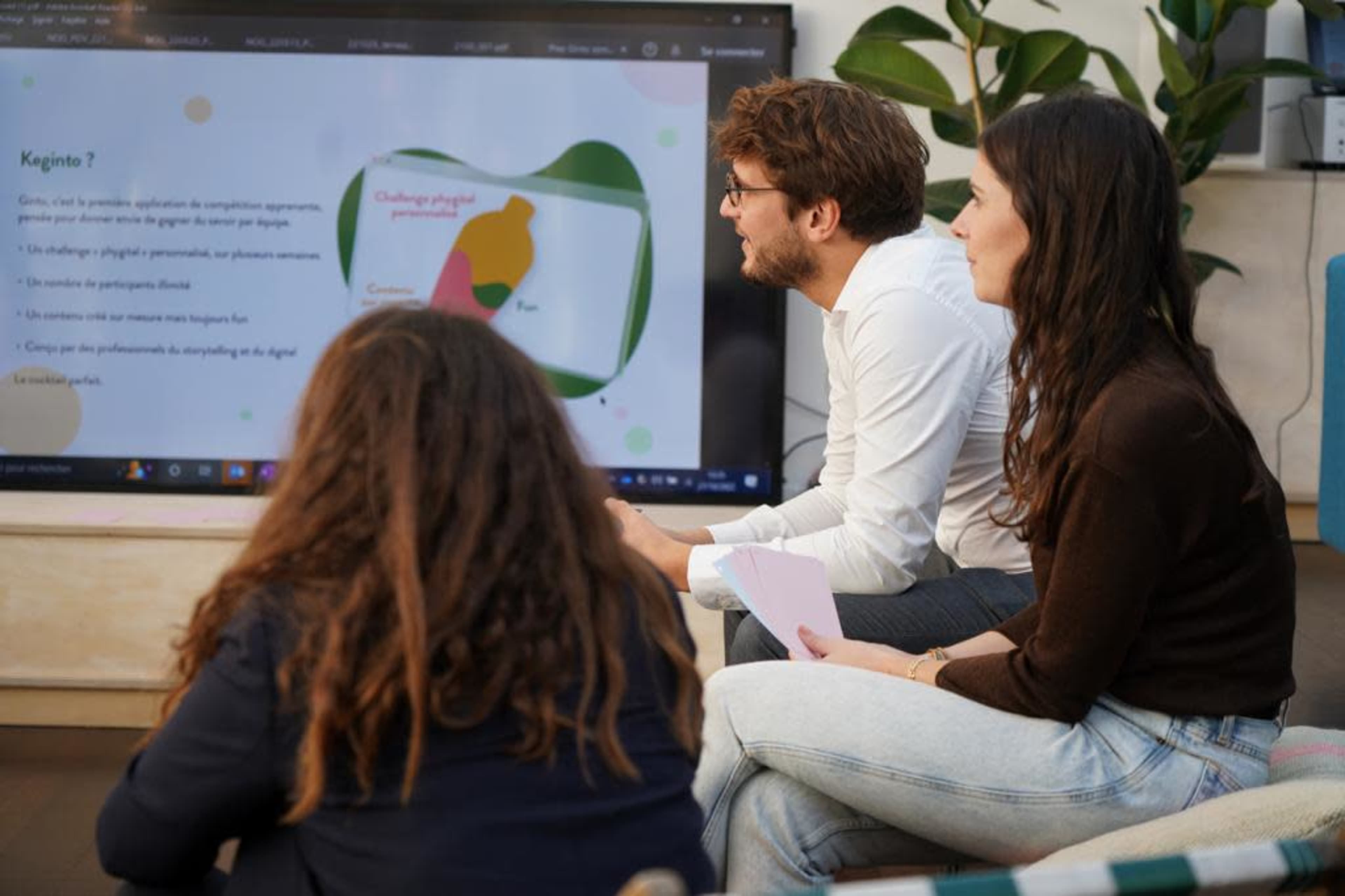 A group of three individuals sits in front of a screen displaying a presentation while discussing a project.