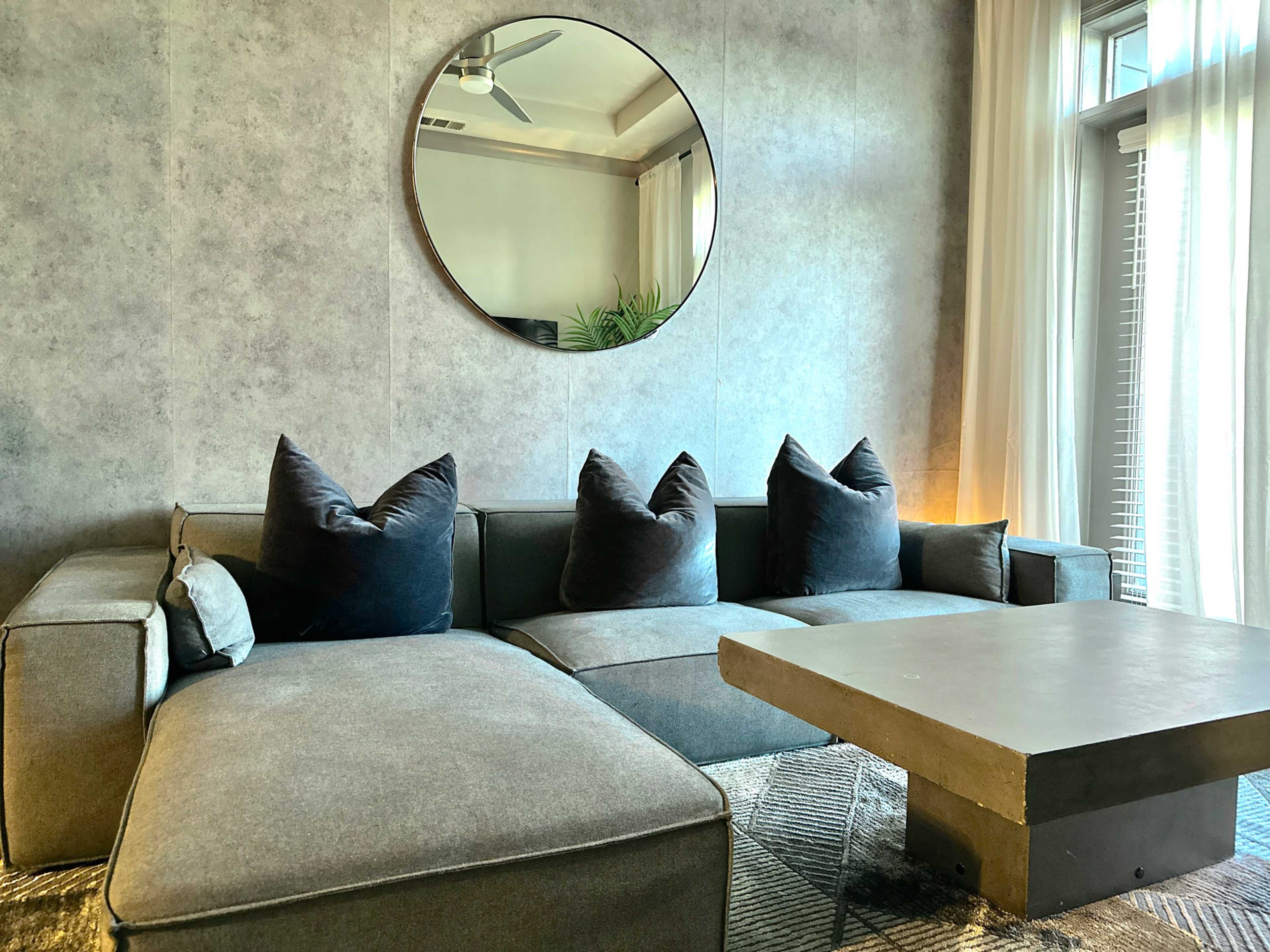A modern living room featuring a gray sectional sofa with large, dark pillows, a concrete coffee table, and a round mirror on the wall.