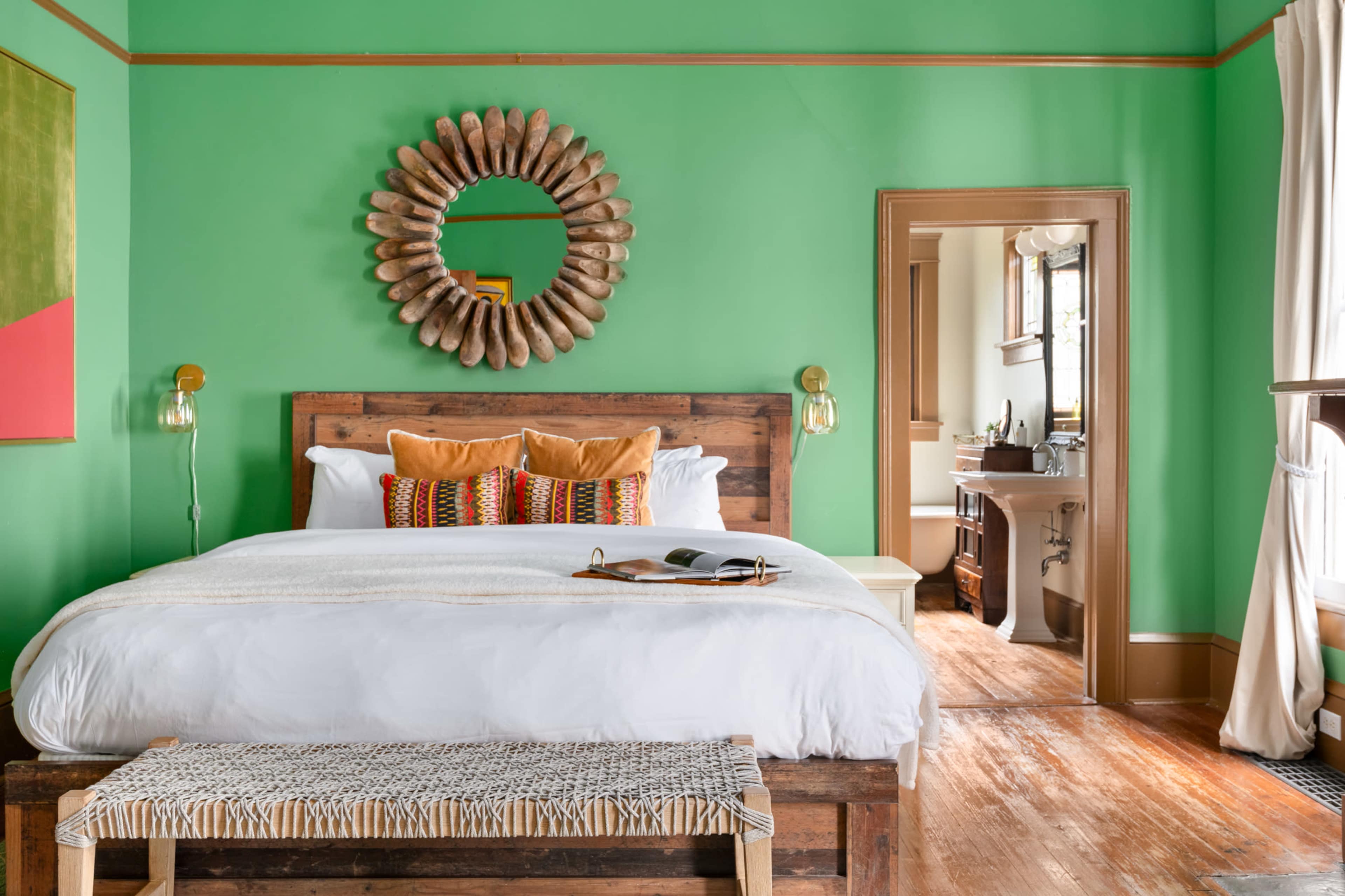 The image shows a brightly colored green bedroom featuring a wooden bed with decorative pillows, a round mirror above the bed, and an adjoining bathroom visible through an open door.