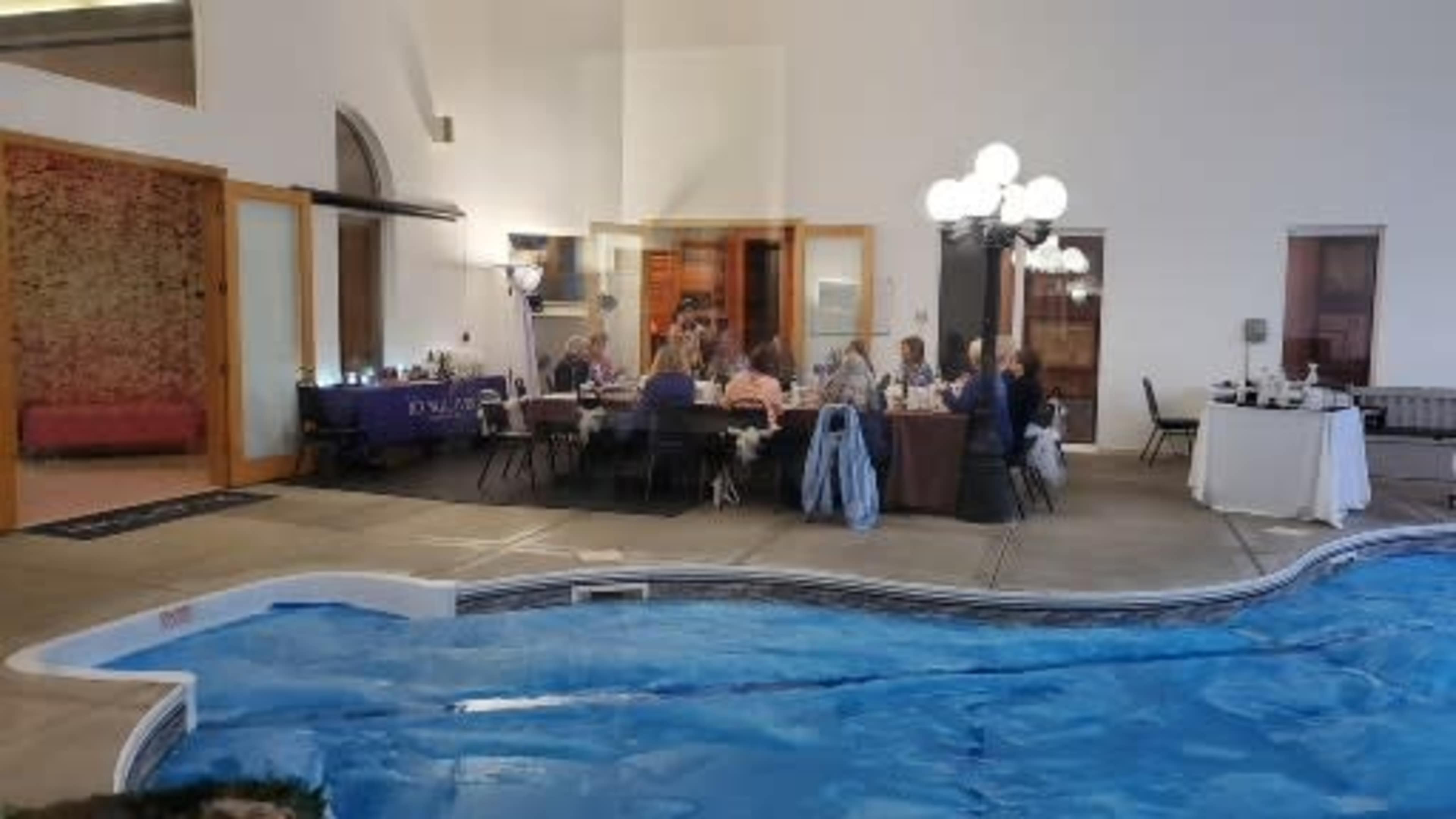A group of people is gathered around a table in a dining area adjacent to a swimming pool.