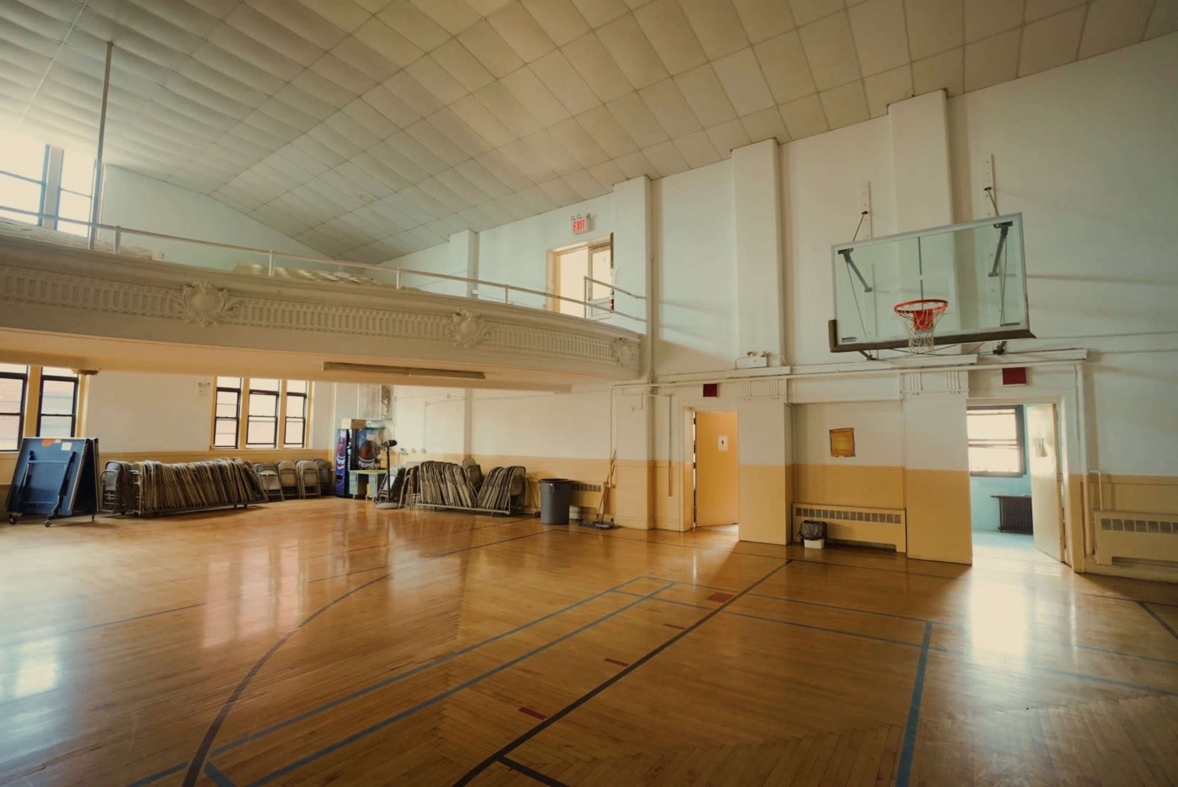 A spacious gymnasium with a basketball hoop, wooden flooring, and stacked chairs against a wall.