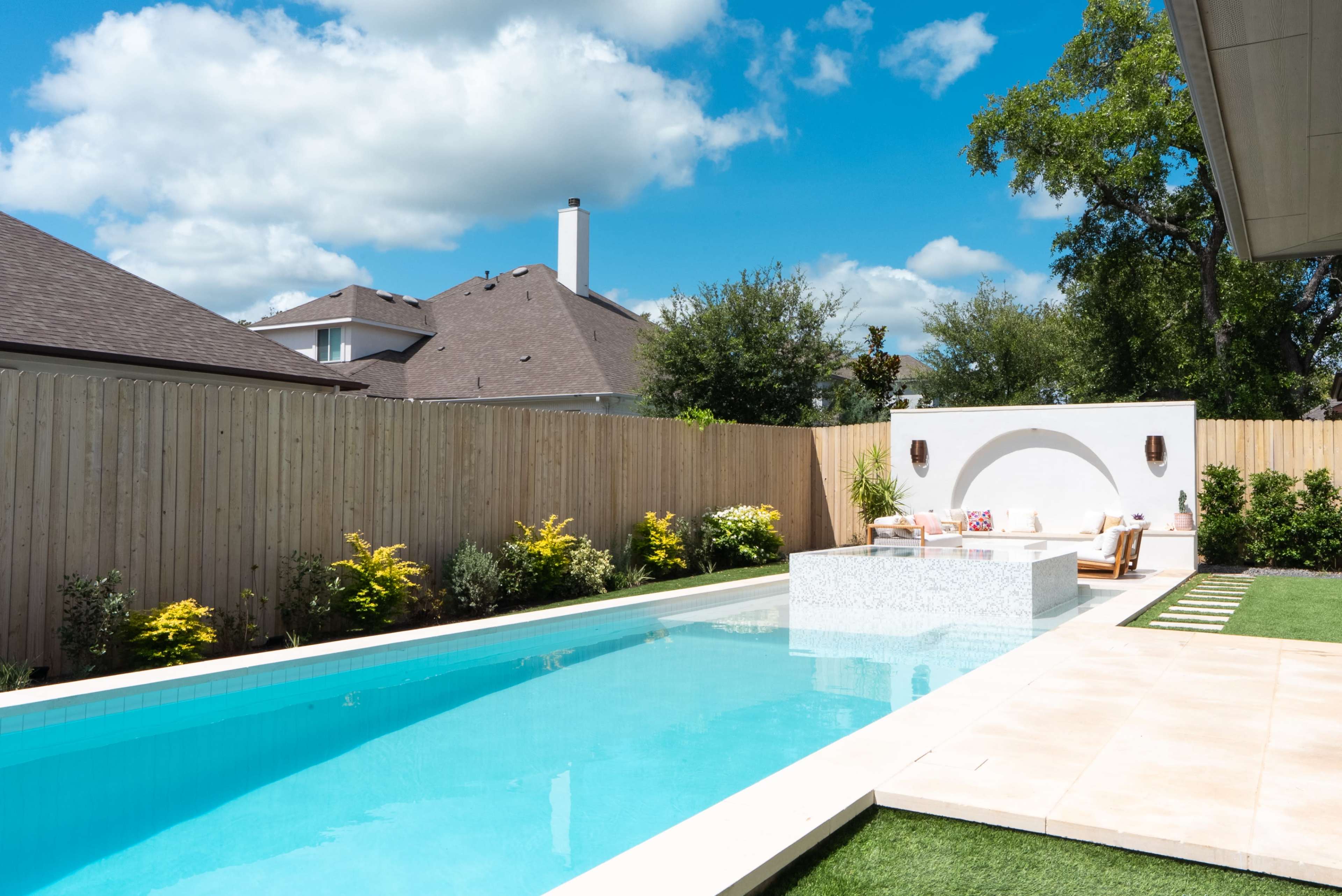 A modern backyard features a rectangular swimming pool with a stone island and lounge chairs, surrounded by greenery and a wooden fence.