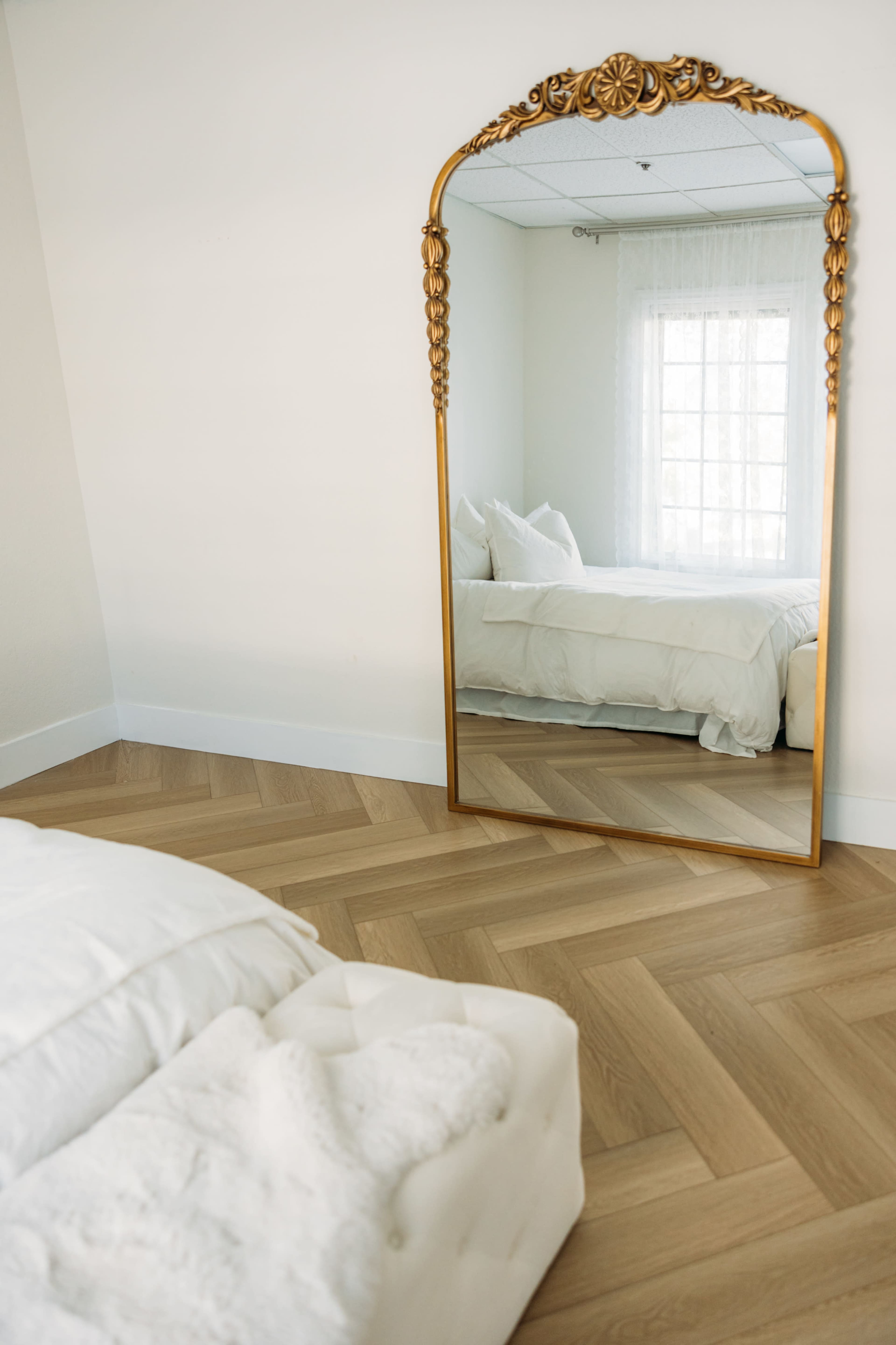 A neatly arranged bedroom with a large ornate mirror reflecting a bed with white bedding, situated on a herringbone-patterned wooden floor.