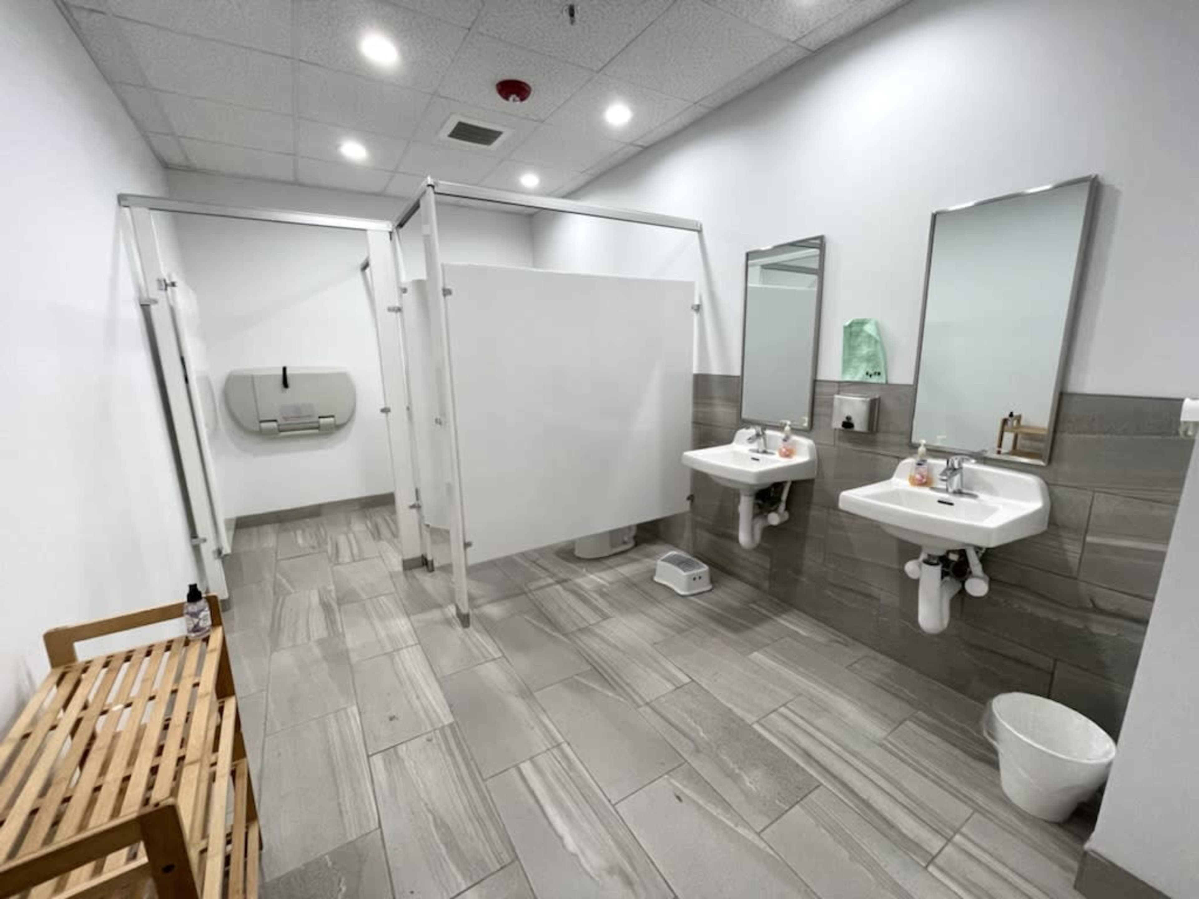 The image shows a clean restroom featuring two sinks, three shower stalls, a wooden bench, and a small wastebasket.