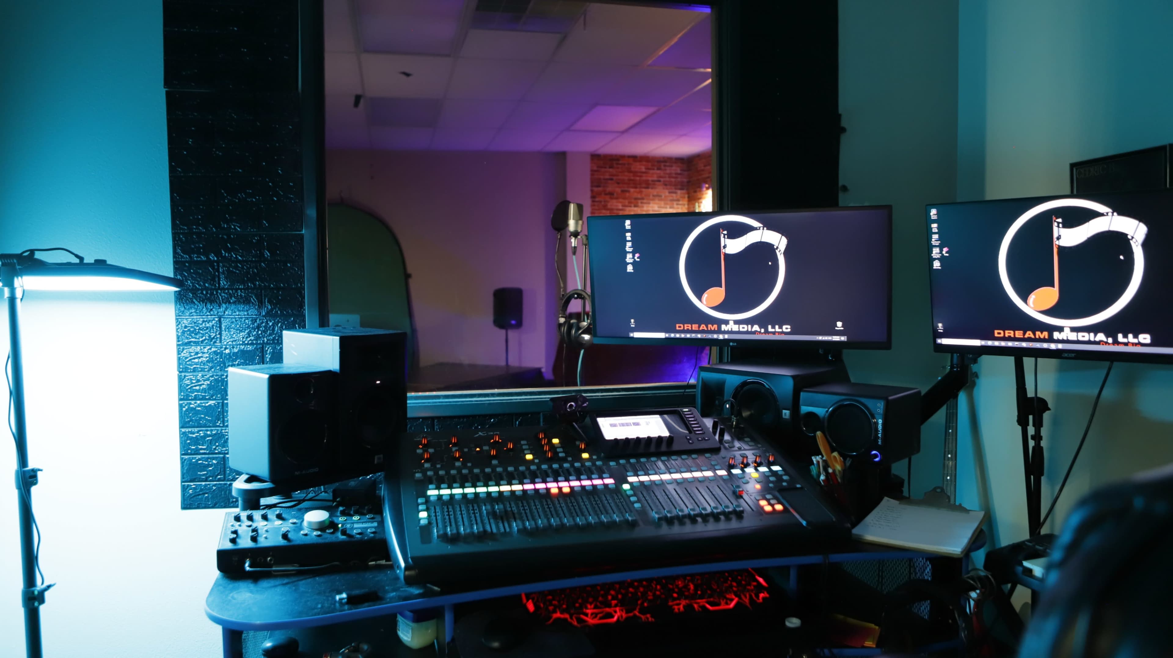 The image shows a sound studio setup with a mixing console, dual computer monitors displaying a logo, and ambient lighting.