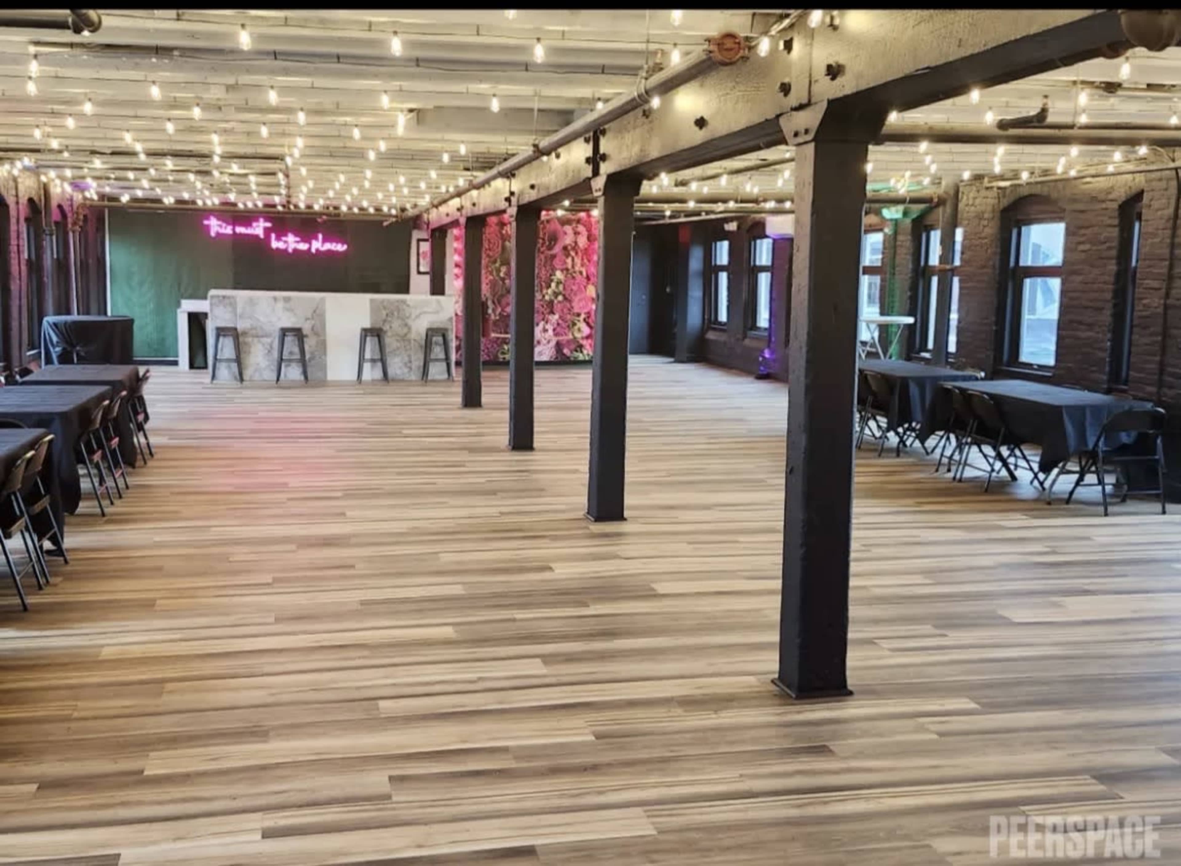 The image shows a spacious event venue with wooden floors, exposed beams, and tables set up in rows, illuminated by string lights and featuring a neon sign.