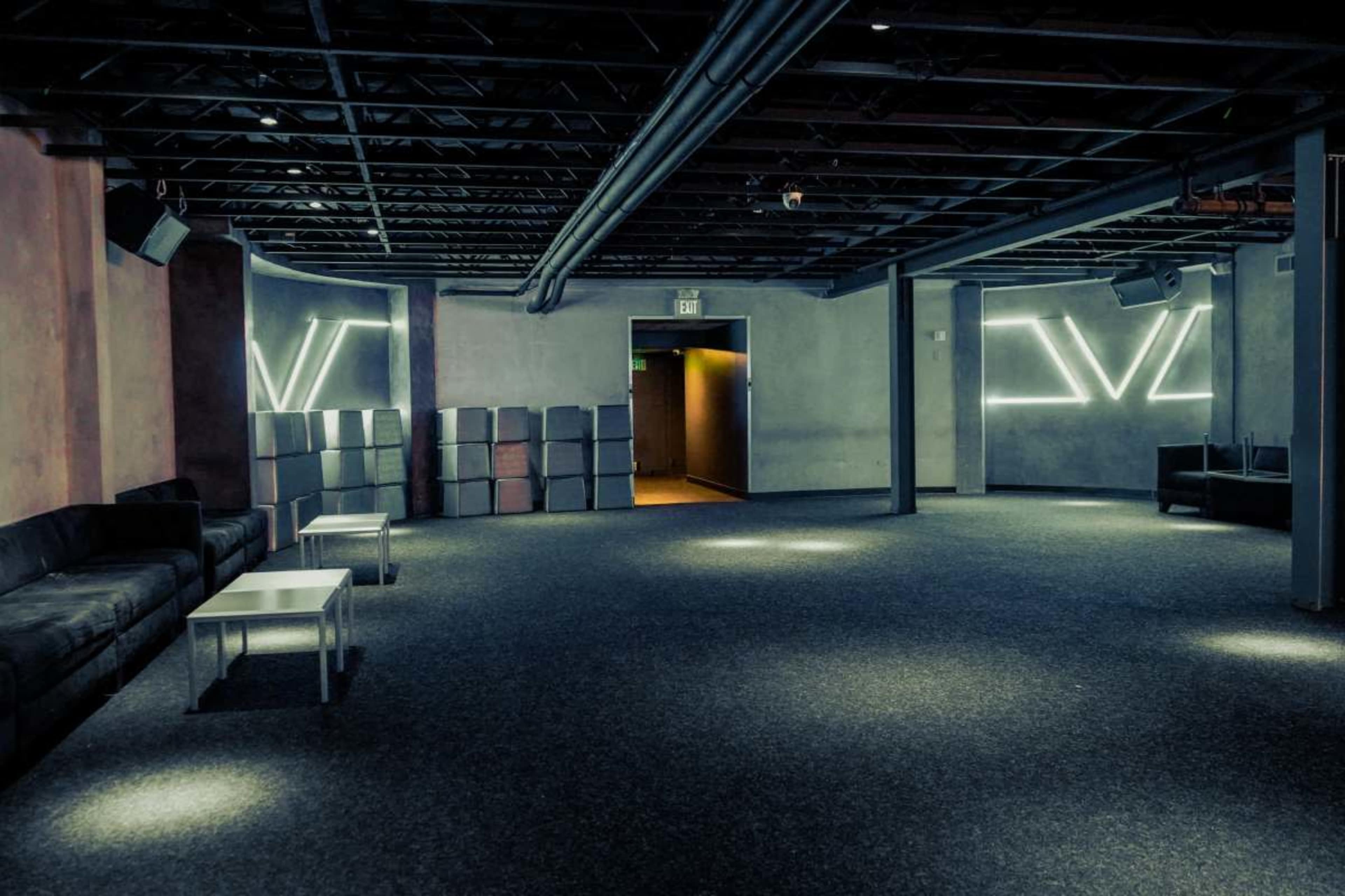 The image shows a spacious, dimly lit room with gray carpet, a few light fixtures, and minimalist furniture, including couches and tables, along with a doorway leading to another area.
