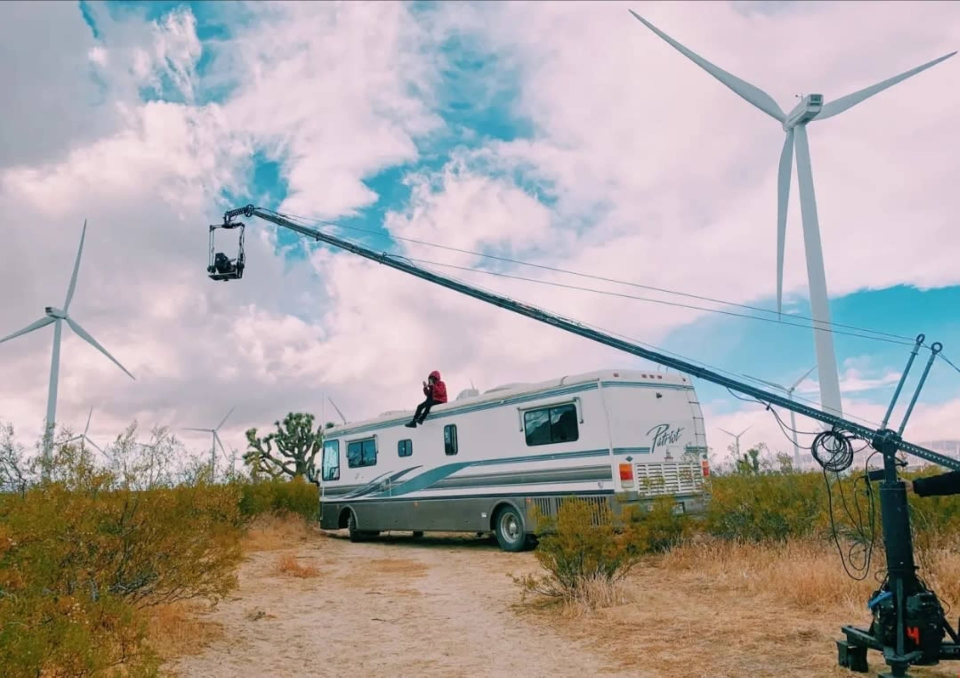 A large RV parked on a dirt path is surrounded by wind turbines and a camera mounted on a crane extends towards the vehicle.