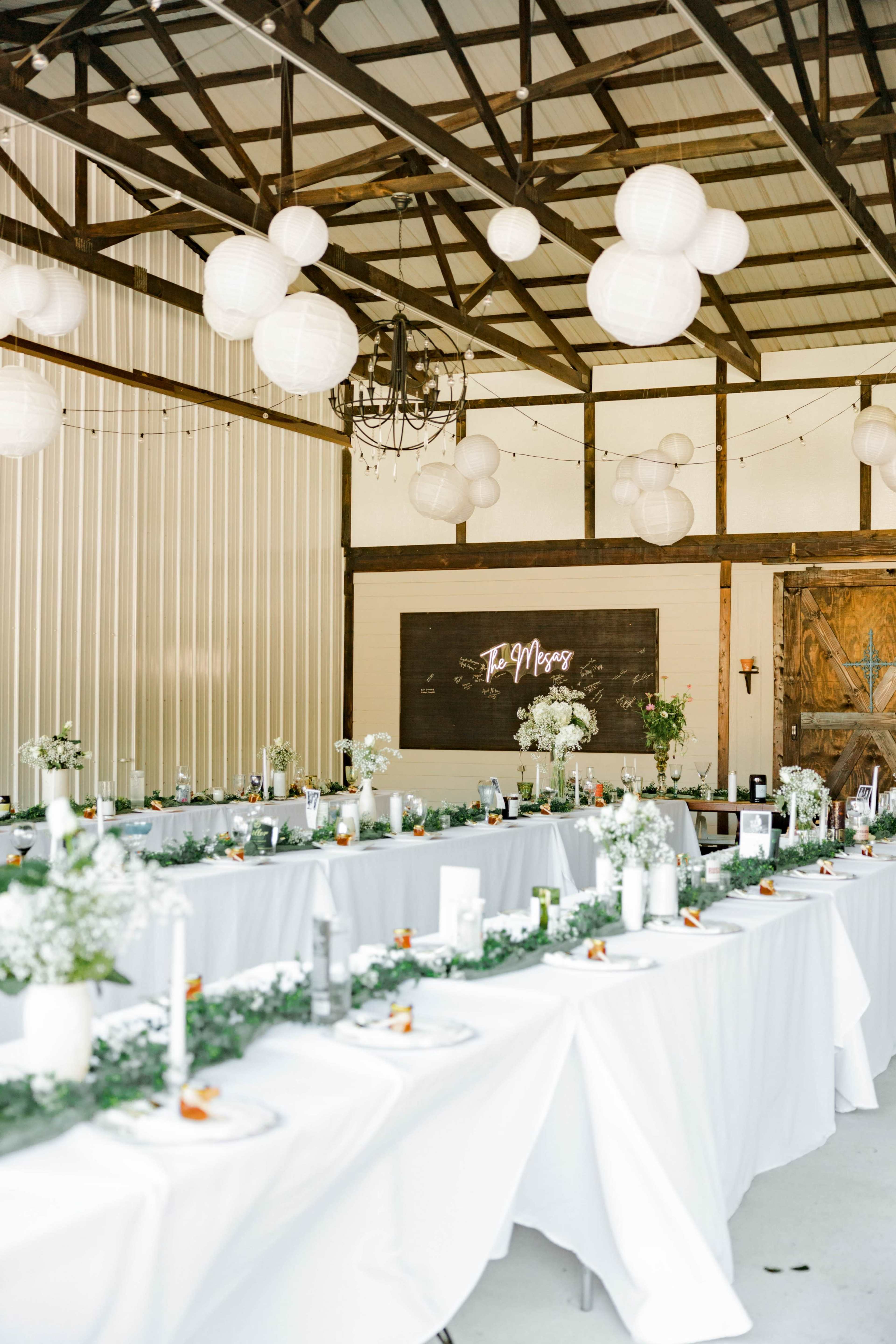A large indoor event space with tables set for a gathering, decorated with white lanterns and floral arrangements.
