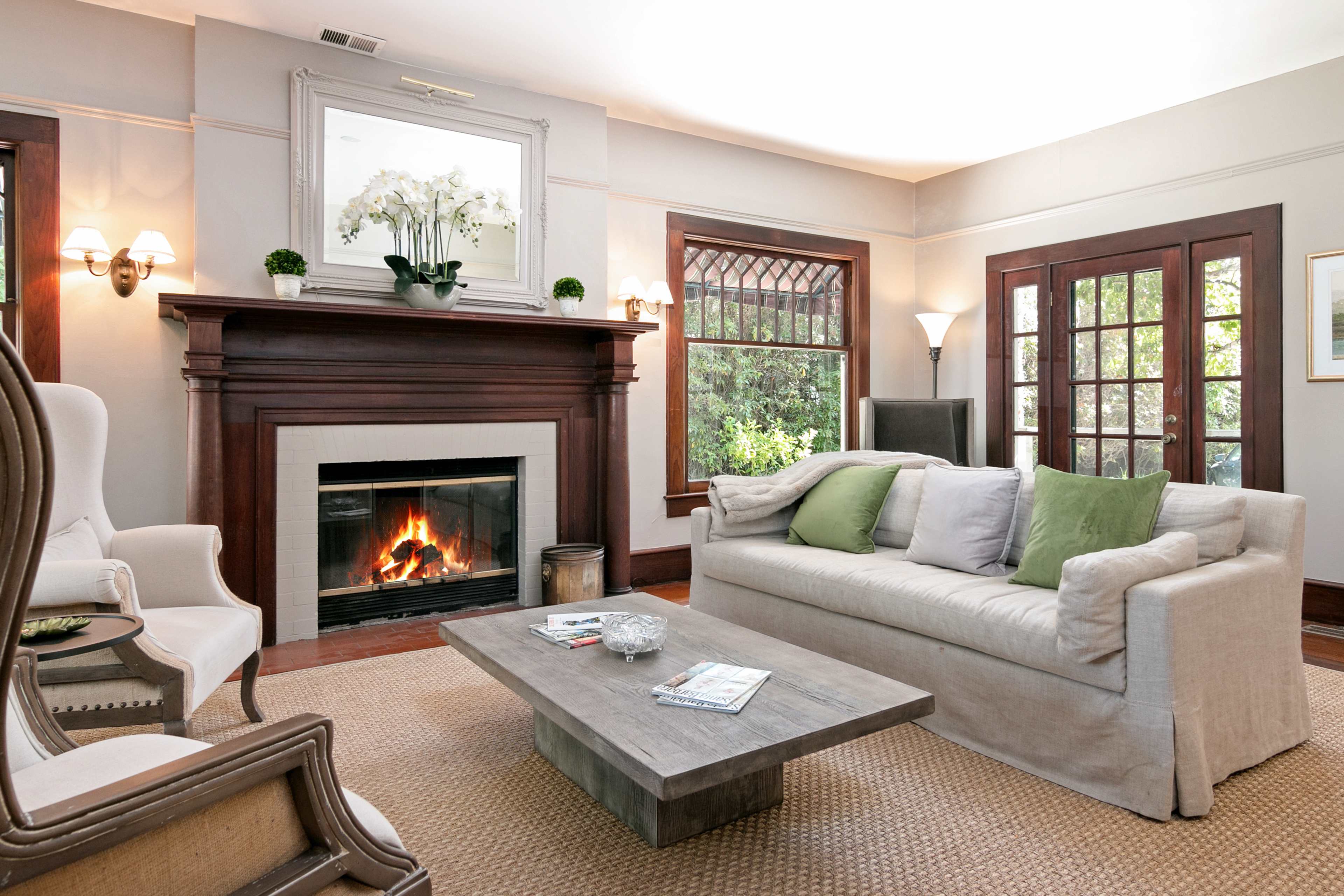 The living room features a light-colored sofa with green accent pillows, a coffee table, and a fireplace framed by a mirrored mantel, surrounded by large windows.