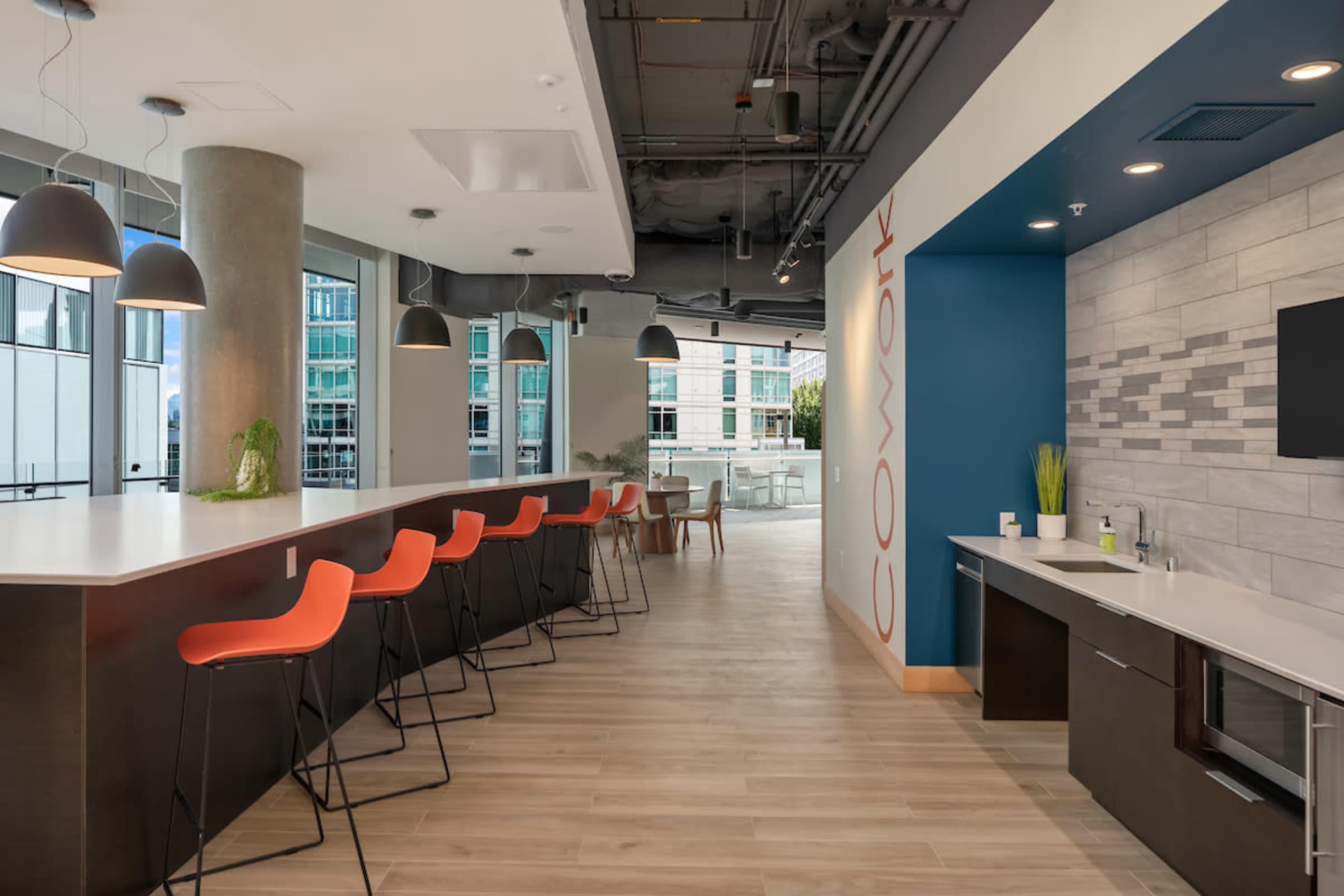 A modern coworking space with a long bar-style table, high stools, and a functional kitchen area.