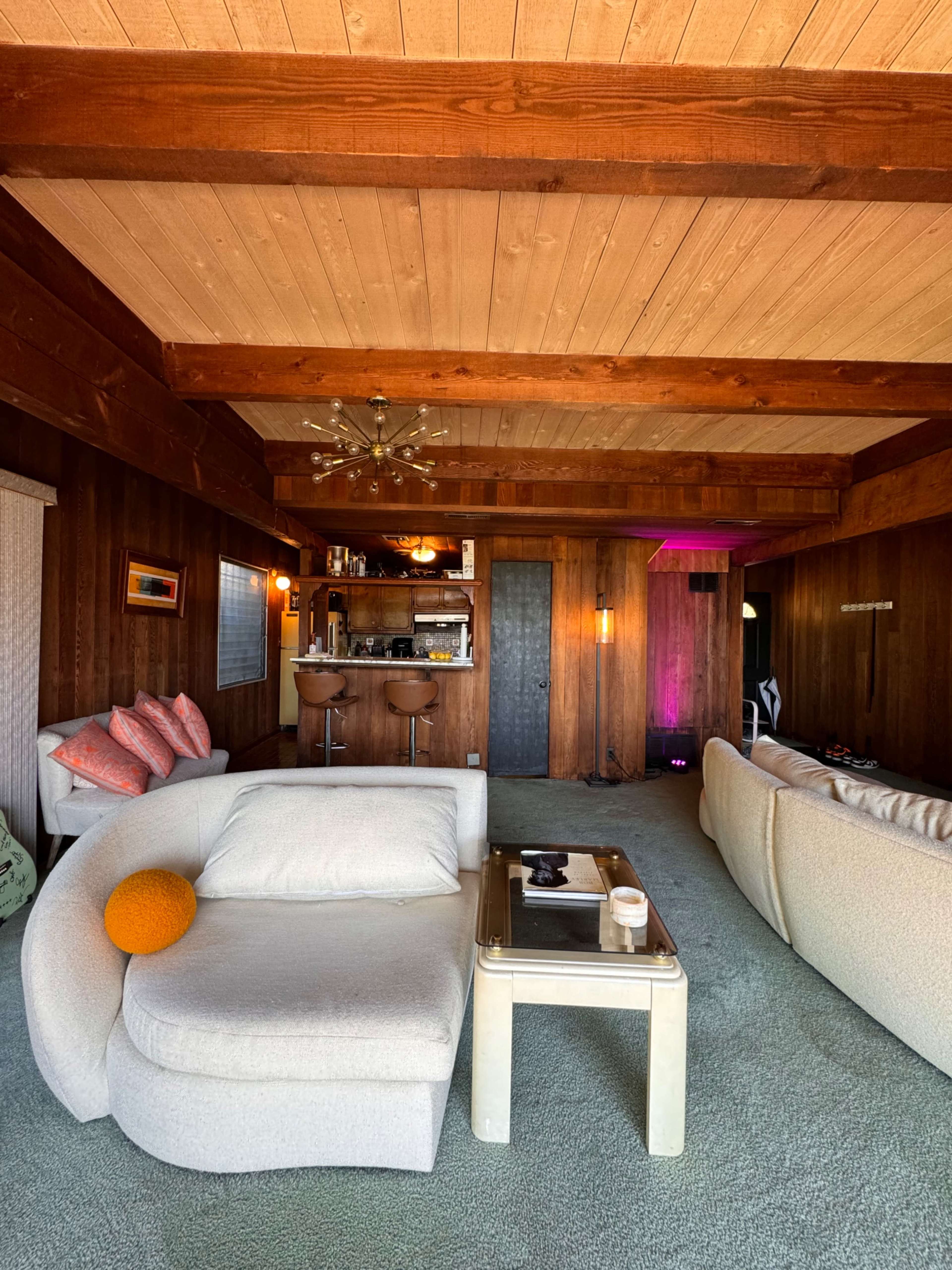 A cozy living room featuring a curved white sofa, a glass coffee table, a bar area in the background, and wooden beams on the ceiling.