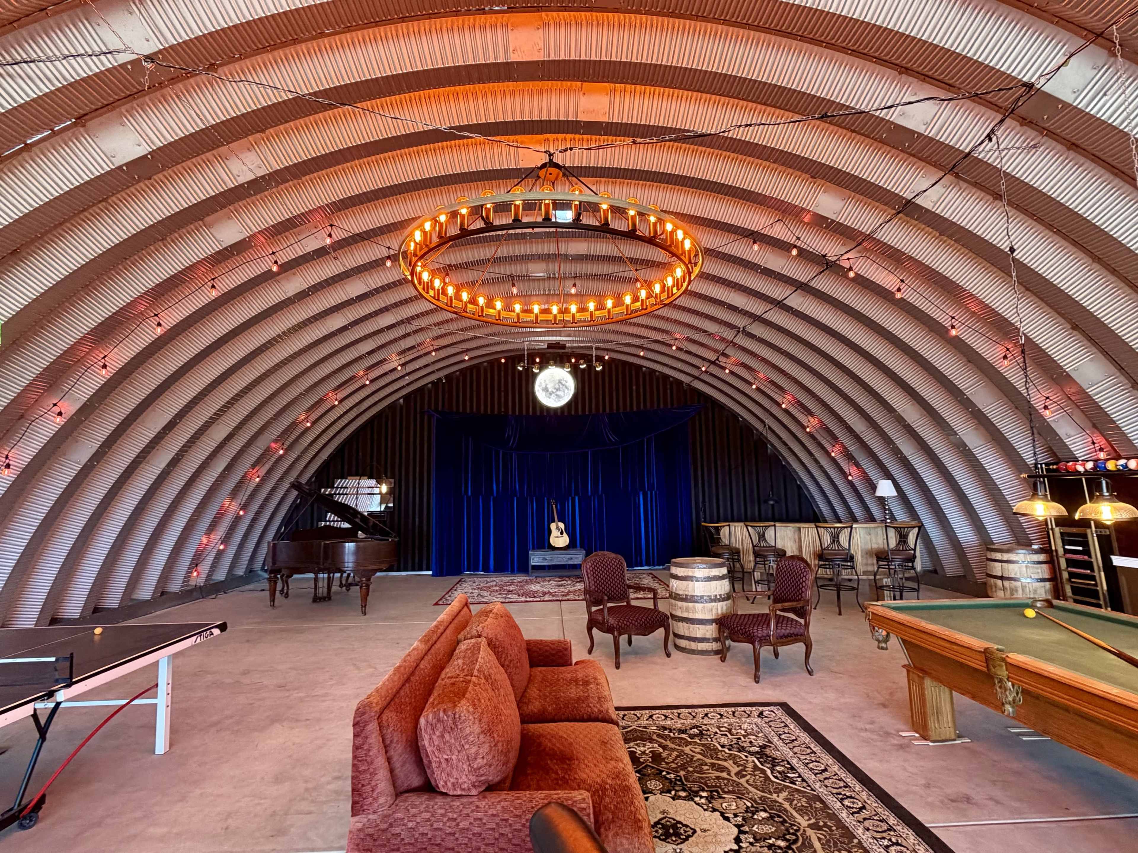 The image shows a spacious, curved room with wooden beams, featuring a piano, seating areas, and decorative lighting elements.
