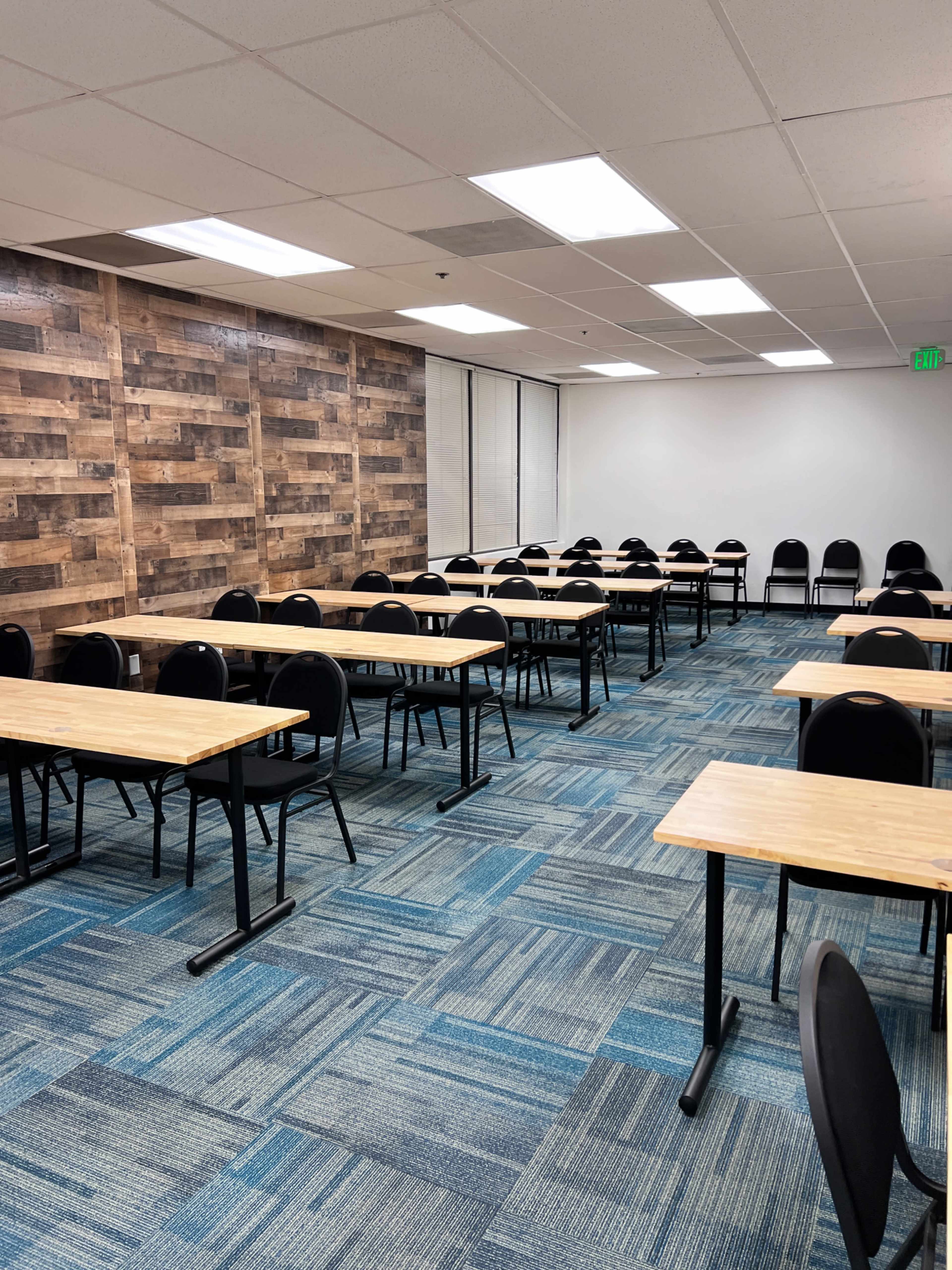 A spacious classroom arranged with rows of wooden tables and black chairs on a blue carpet.
