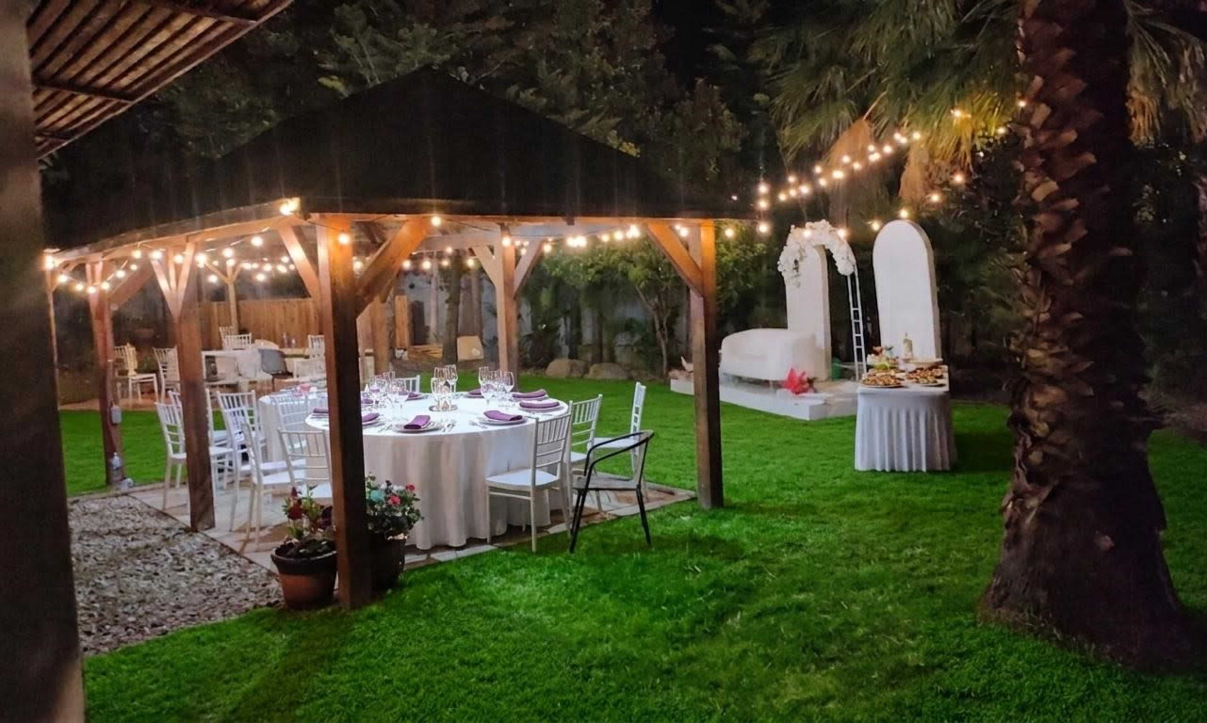A decorated outdoor dining area features a round table set with white linens, surrounded by chairs, under a gazebo illuminated by string lights.