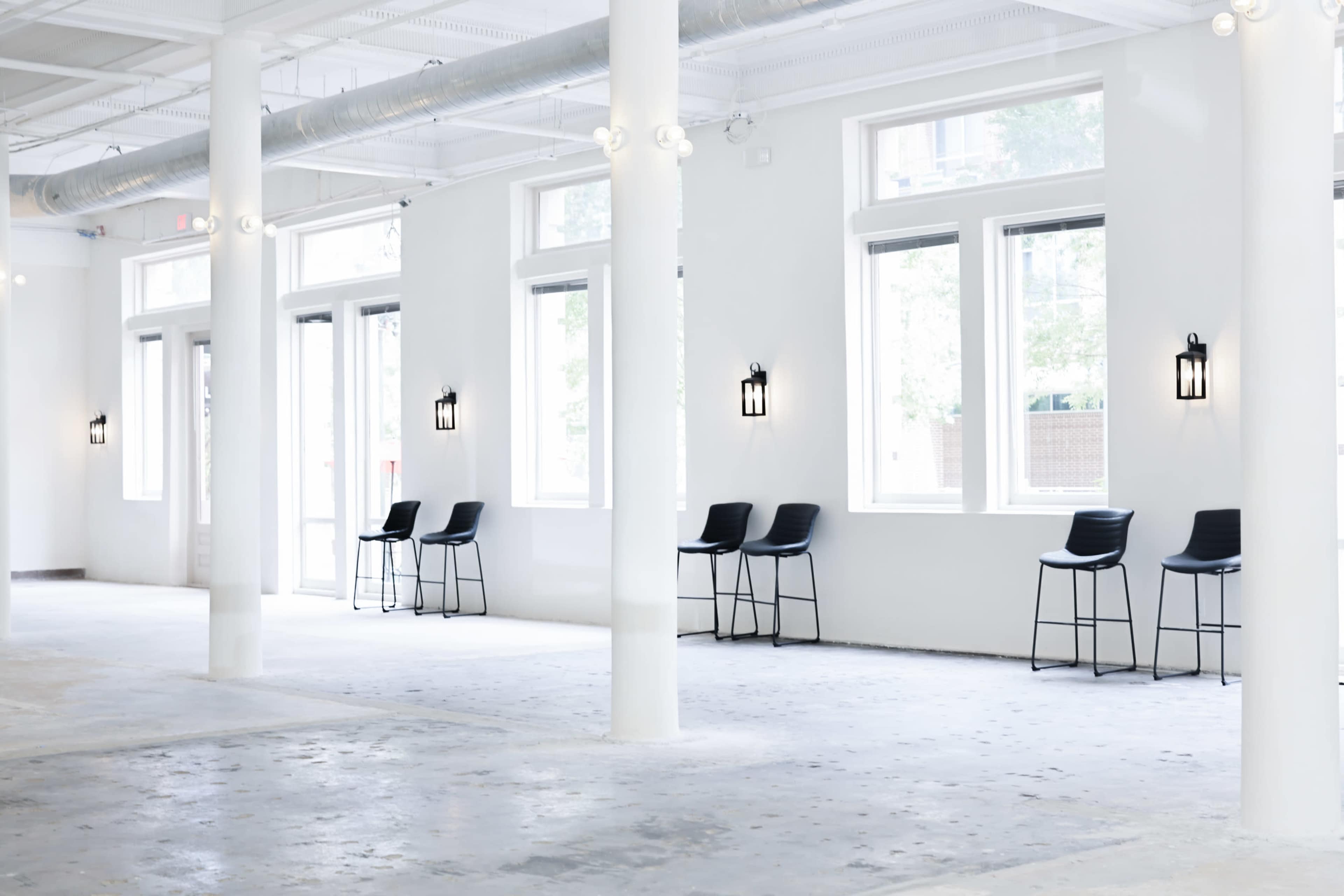 The image shows a spacious, empty room with white walls, several large windows, and a row of black bar stools along the side.