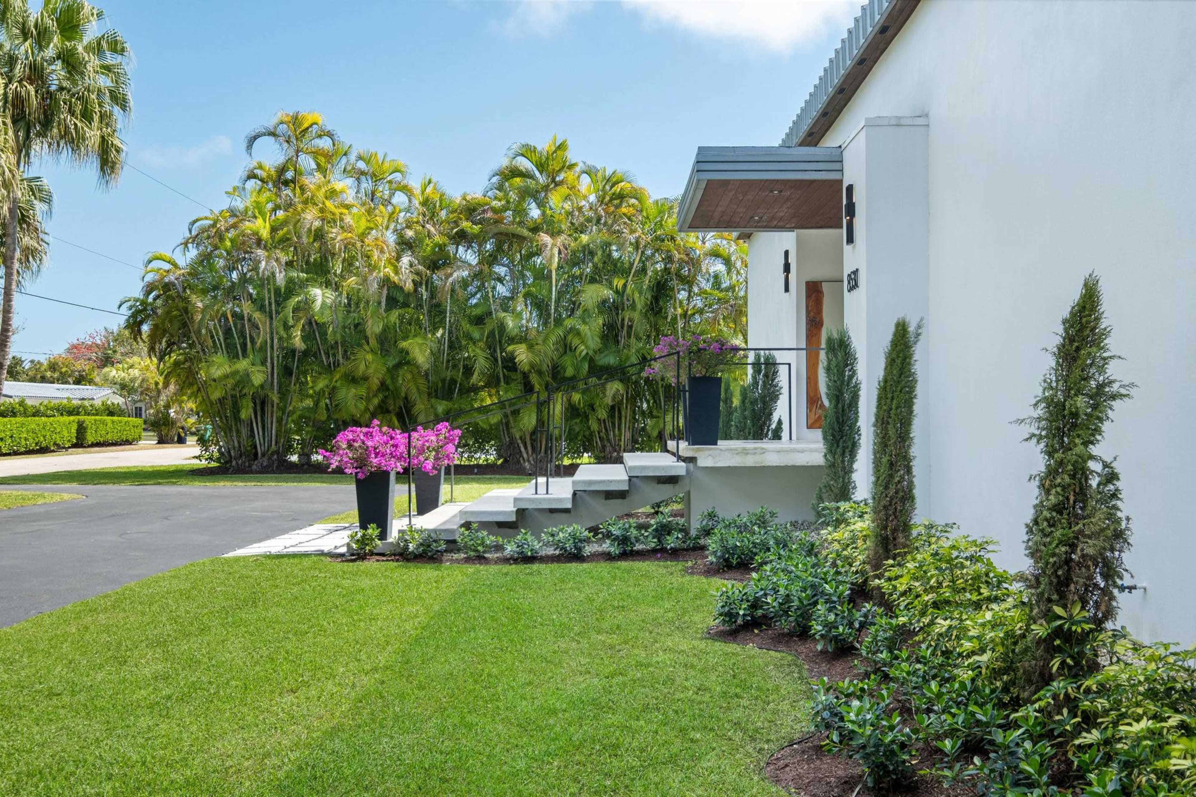 A modern home features a neatly landscaped front yard with green grass, steps leading to the entrance, and tropical plants in the background.