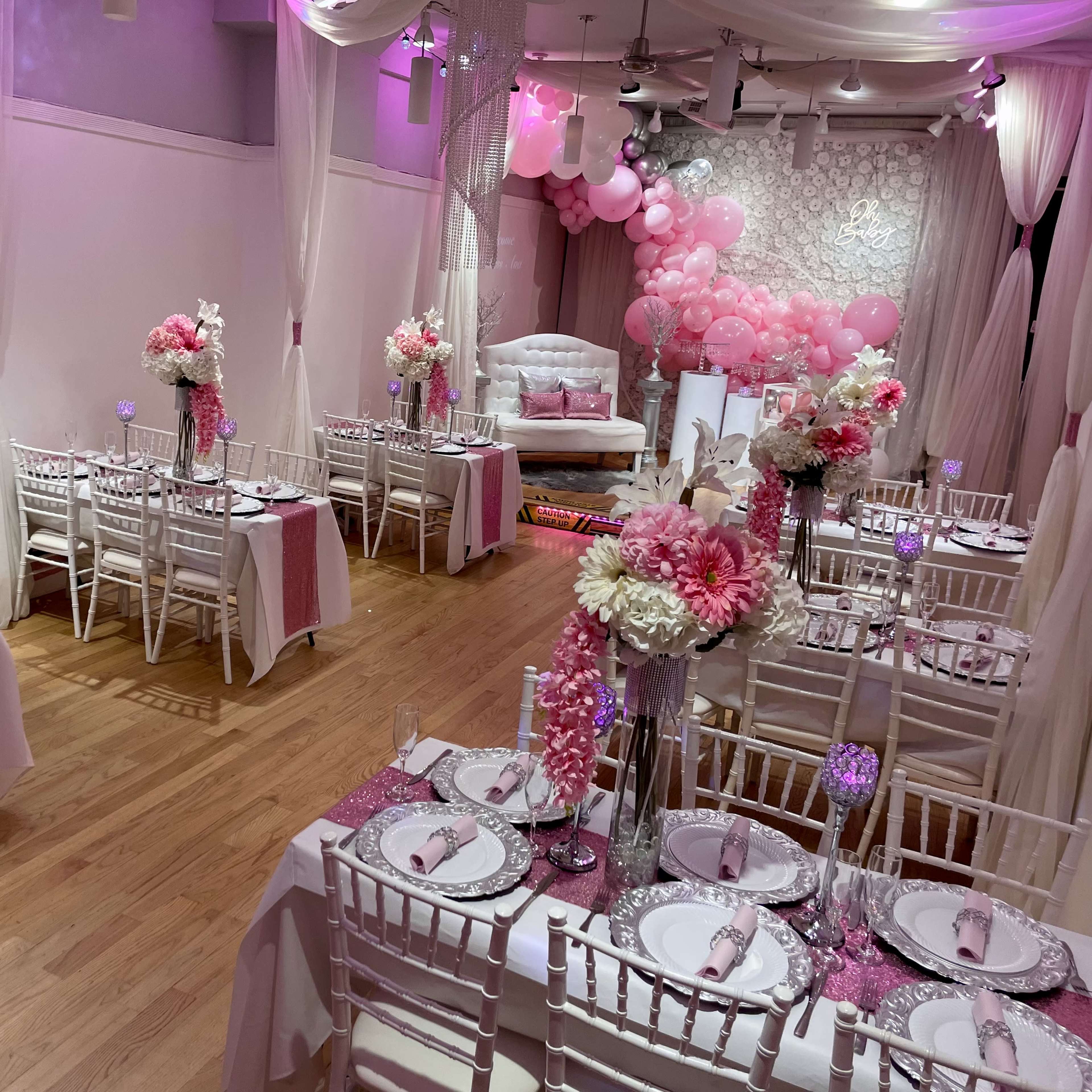A decorated event space features elegantly arranged tables with pink floral centerpieces and balloons, creating a festive atmosphere.