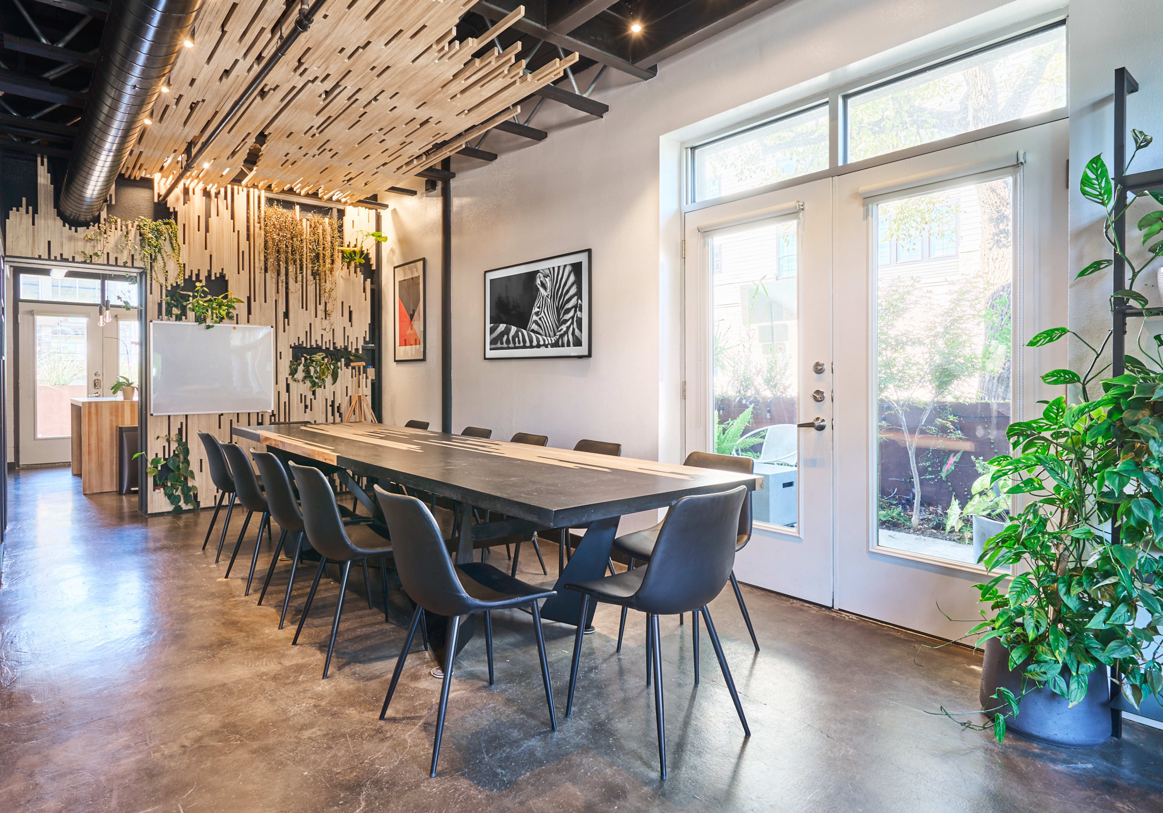 A large conference room features a wooden table surrounded by black chairs, with natural light streaming in through two doorways and decorative plants along the walls.
