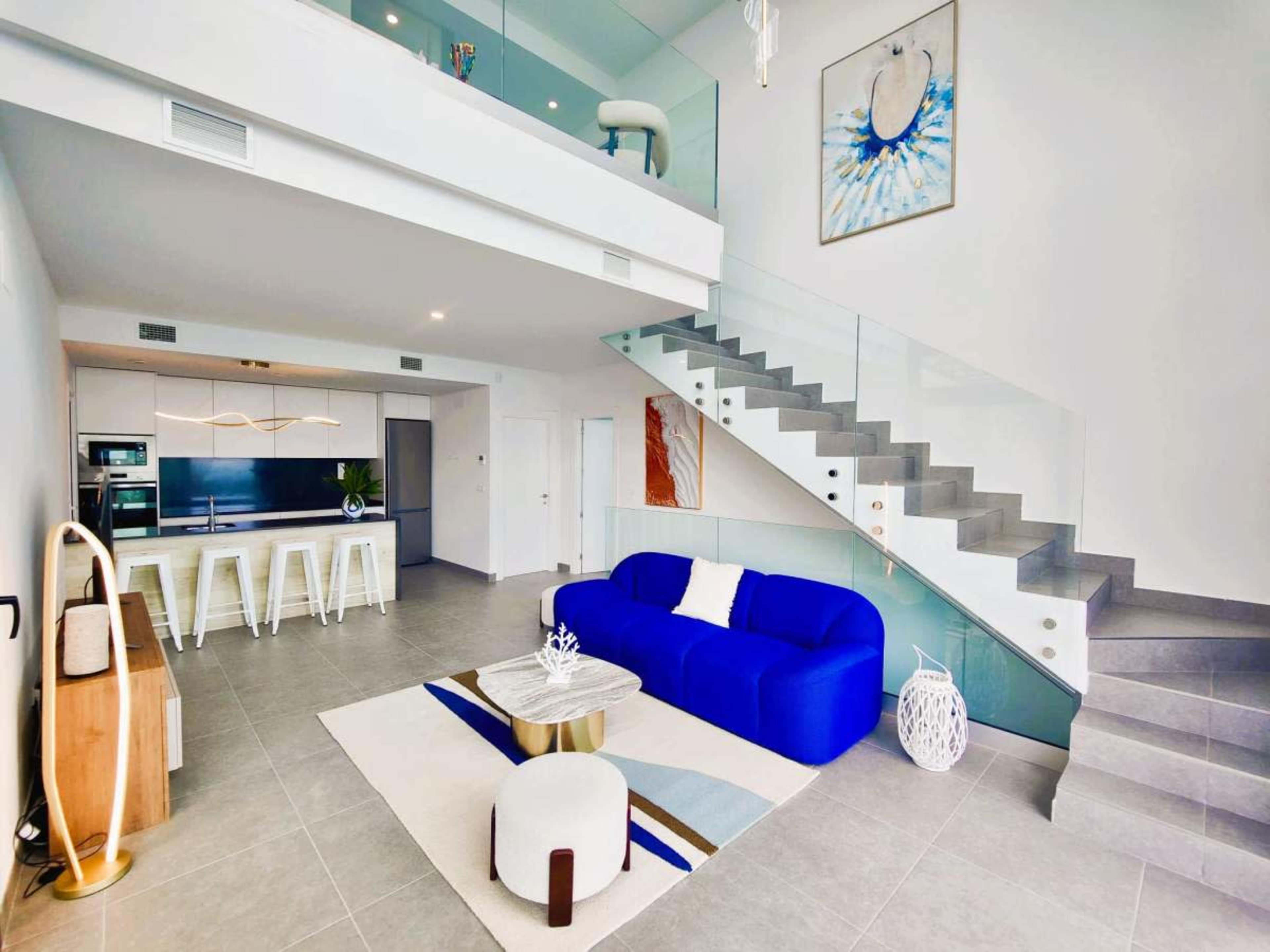 The image shows a modern two-level living space featuring a blue couch, a white round table, and a staircase with glass railings.