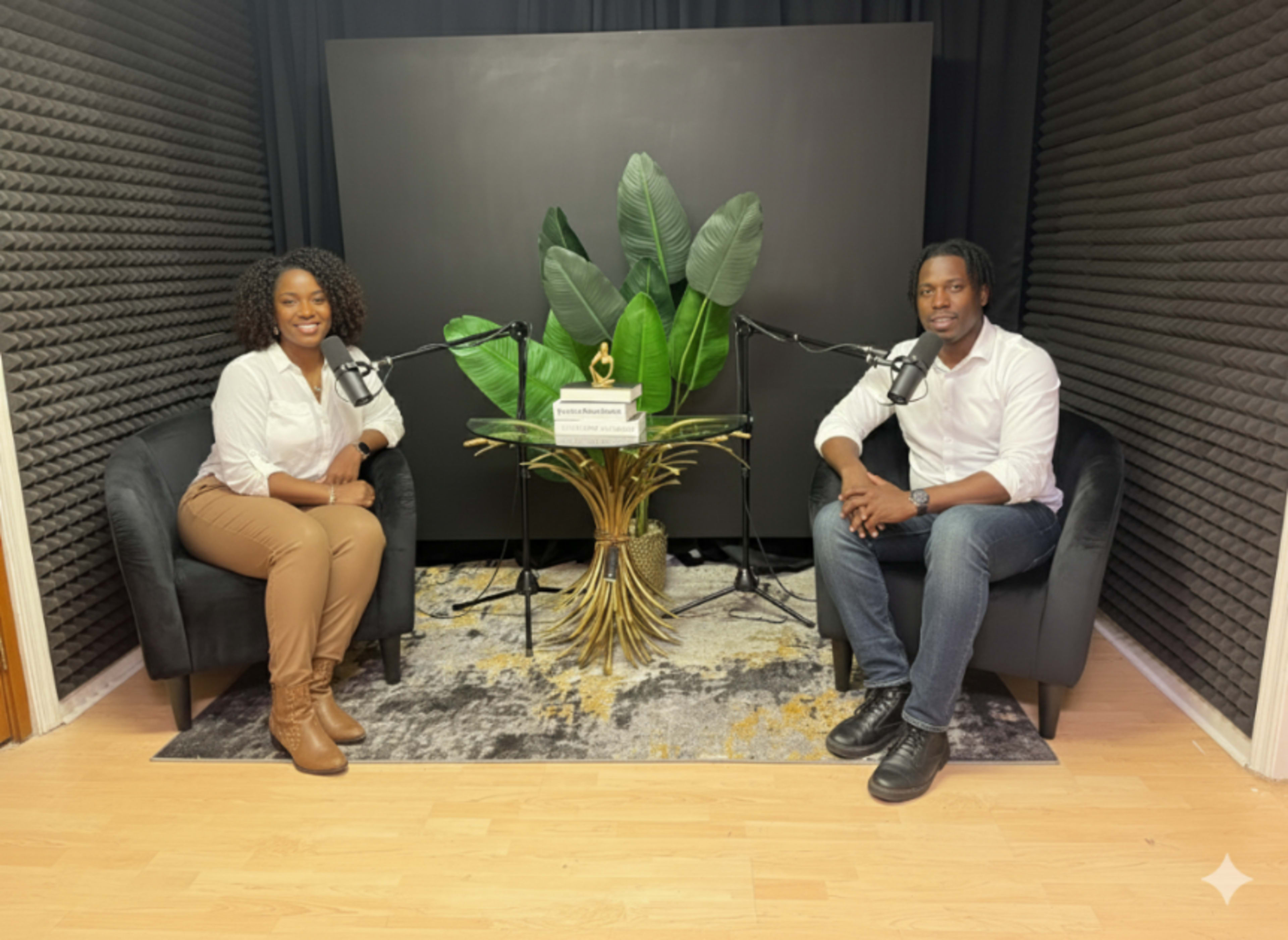 Two people sit on black chairs in a small studio, surrounded by soundproof walls, with a large plant and microphones positioned on a table between them.