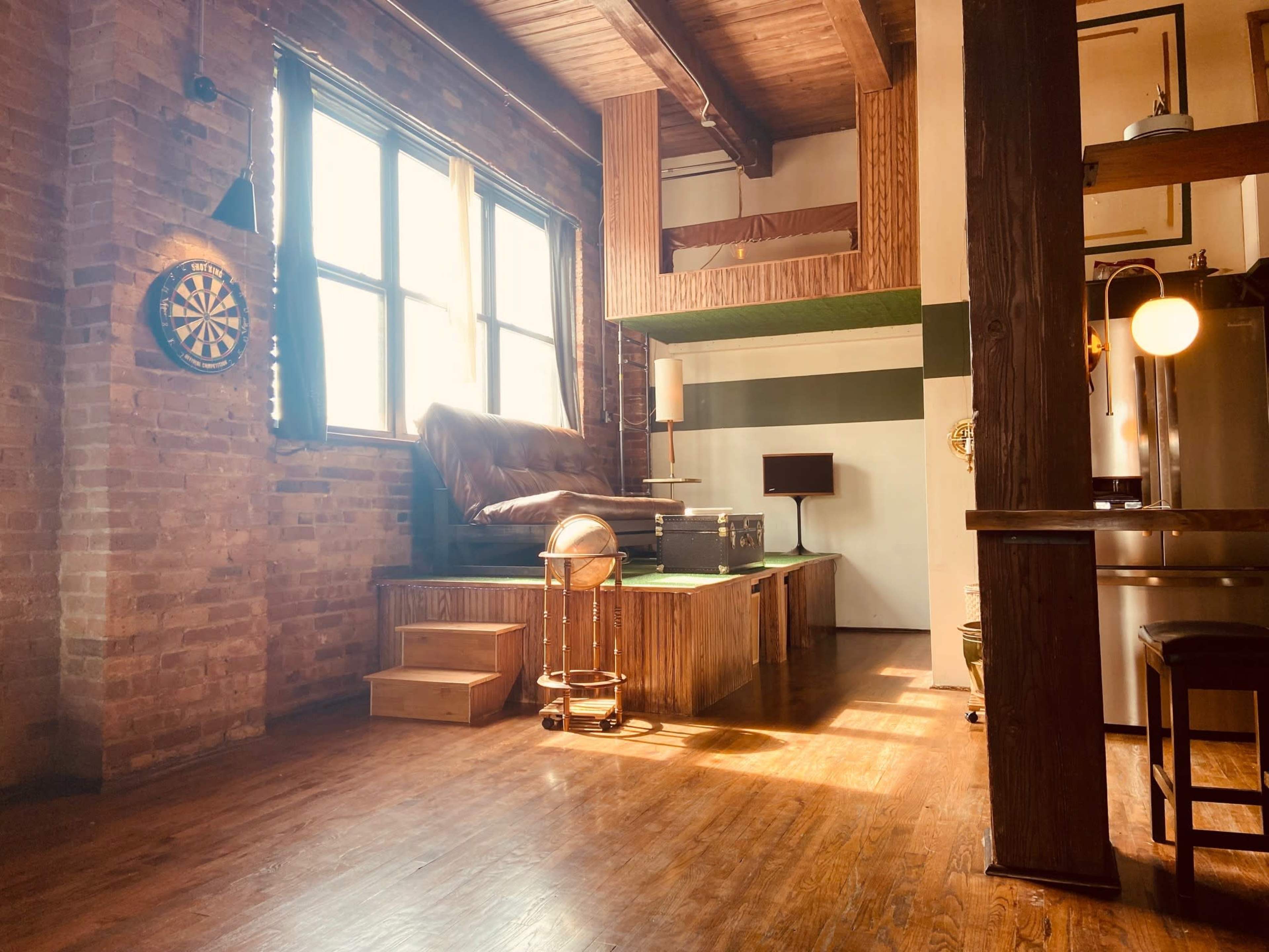 A rustic, open space featuring exposed brick walls, wooden floors, and a lofted area with a bed, darts board, and a small seating area.