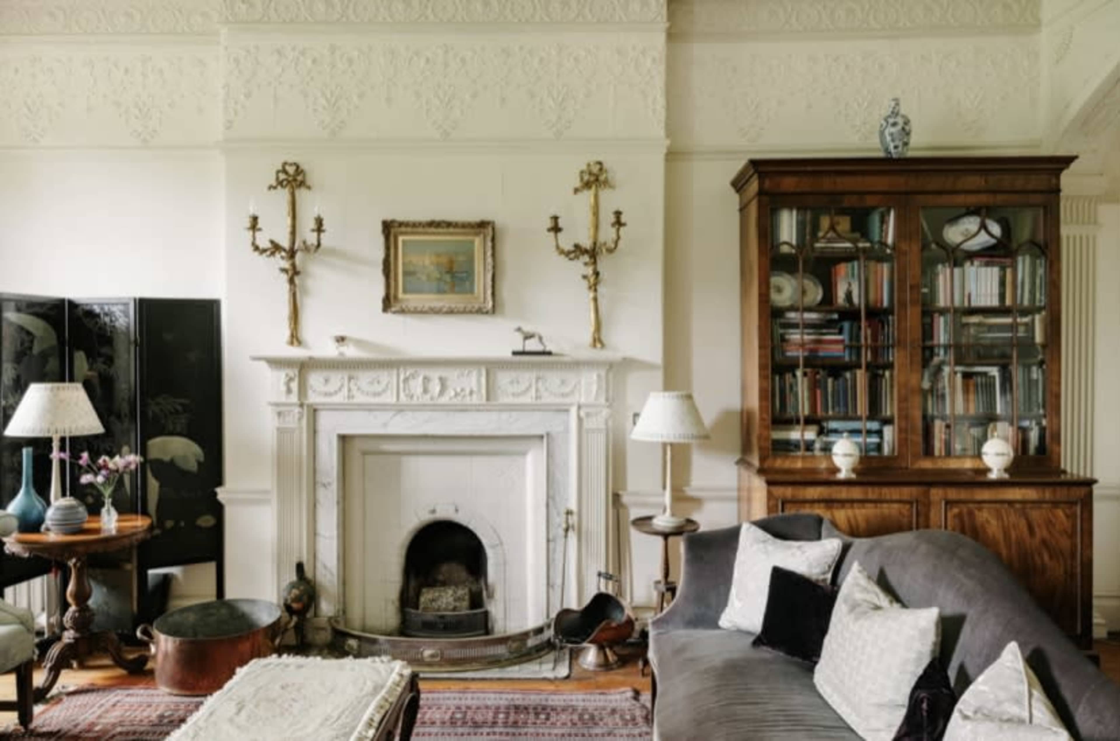 A living room with a fireplace, a wooden cabinet filled with books, and a sofa, accompanied by decorative items on the mantle and tables.