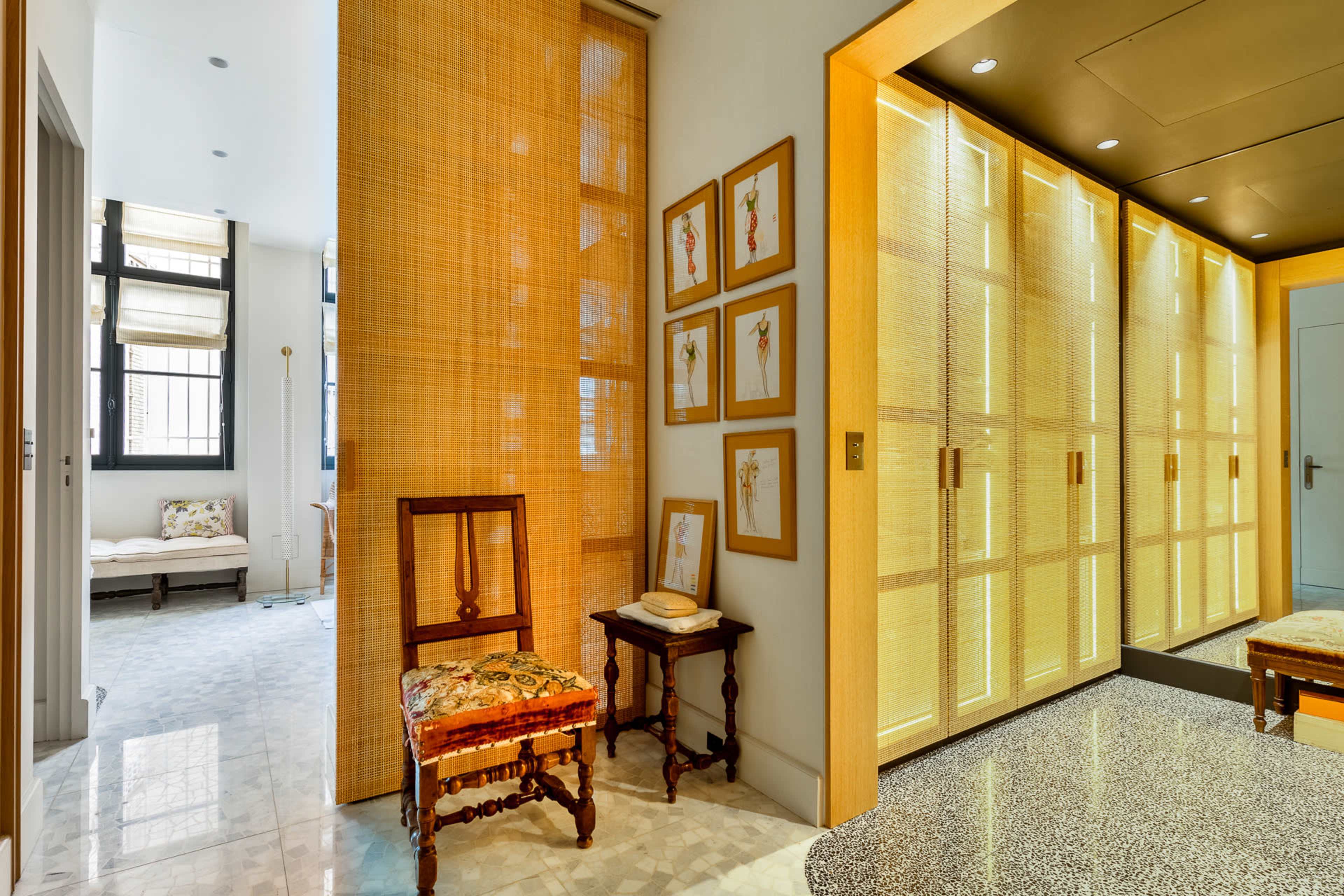 The image shows a hallway with light wood paneling, decorative framed pictures on the wall, and a mirrored closet door, leading to a softly lit living space.