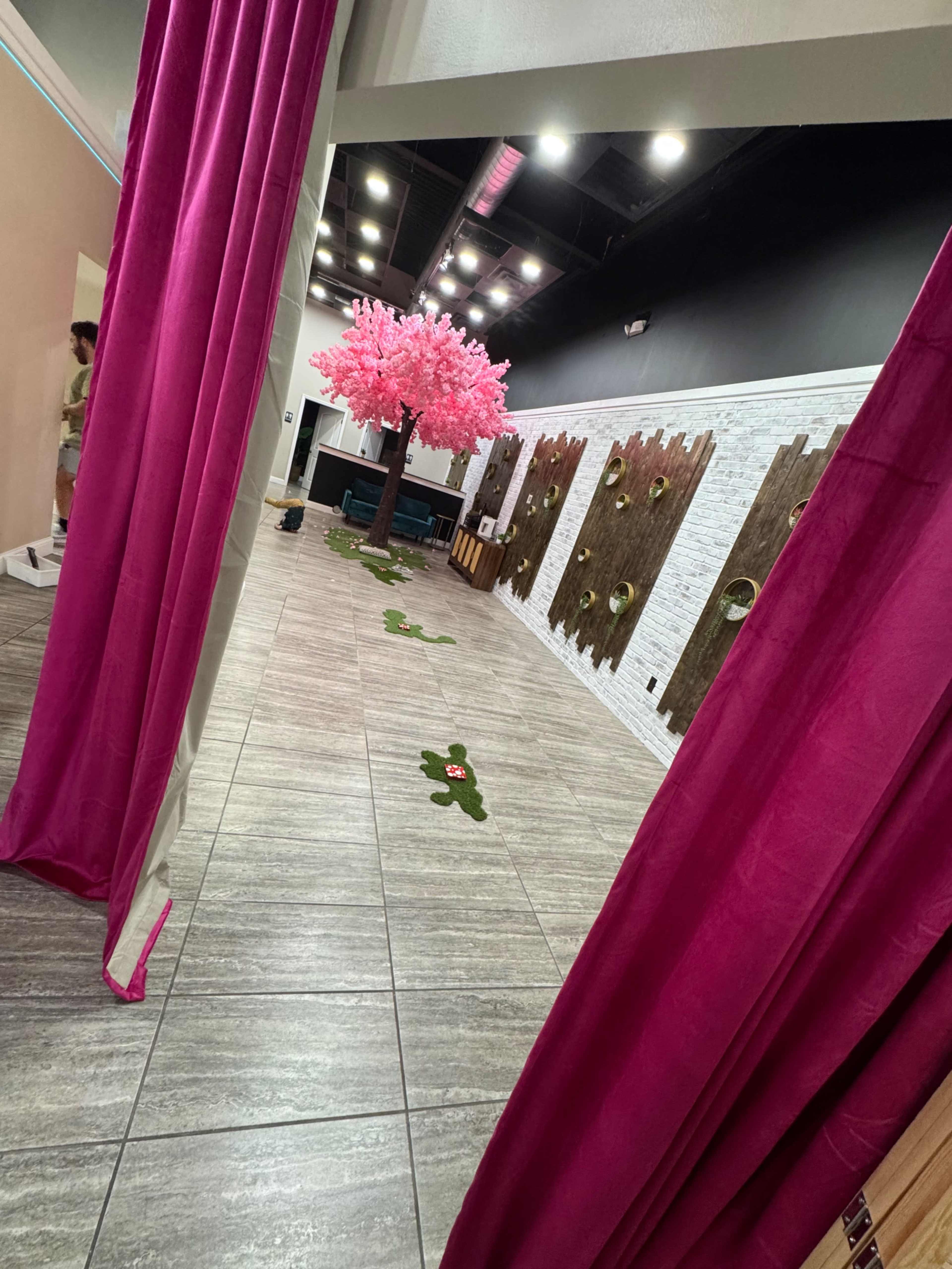 The image shows a brightly lit indoor space with pink curtains, a decorative pink tree, and wooden wall panels along the corridor.