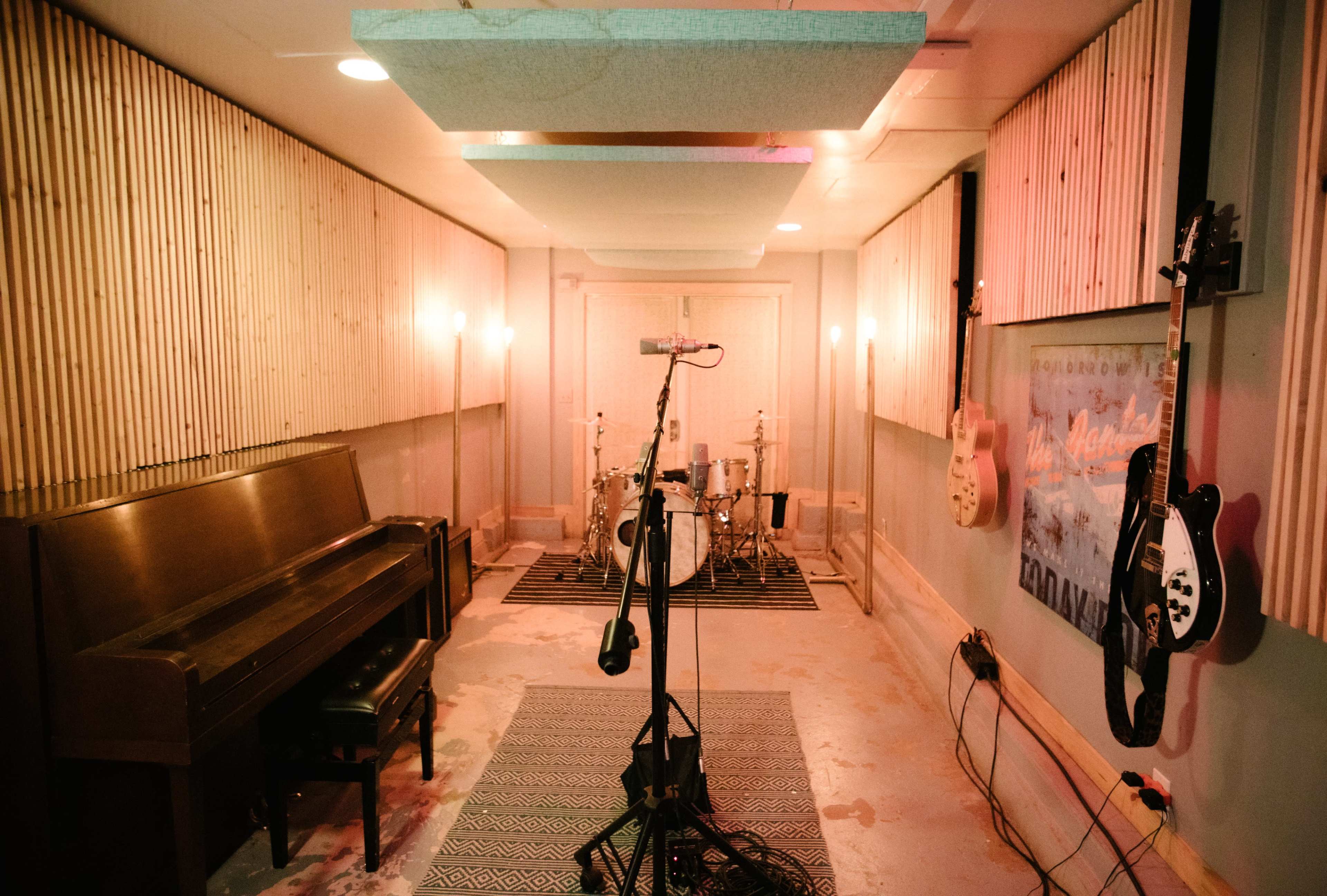 A music recording studio featuring a piano, a drum set, and electric guitars mounted on the wall.