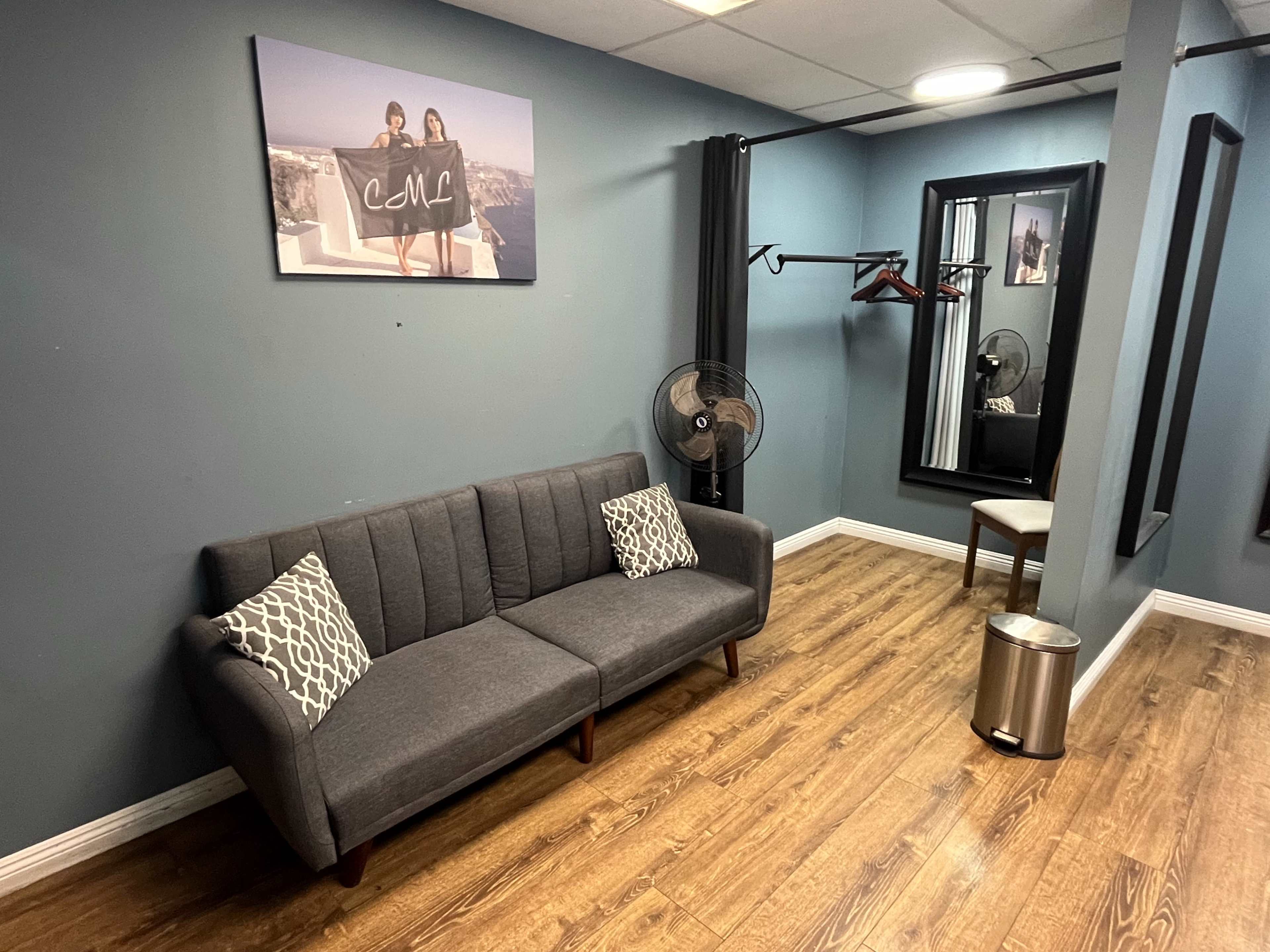 The image shows a small waiting area with a gray sofa, two fans, a mirror, and a trash can against a blue wall.