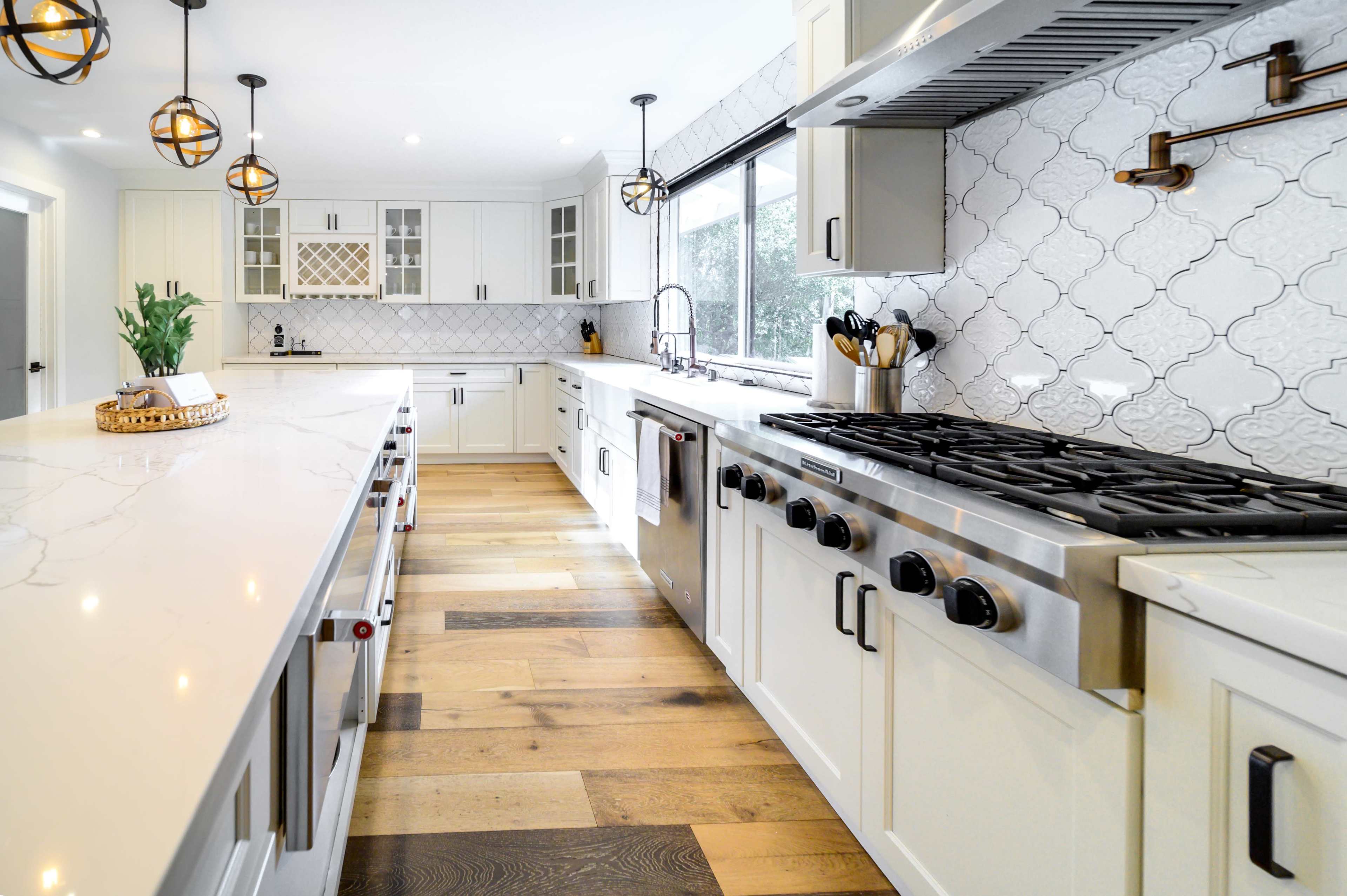 The image shows a modern kitchen with white cabinetry, a large marble countertop, a gas range, and pendant lighting.