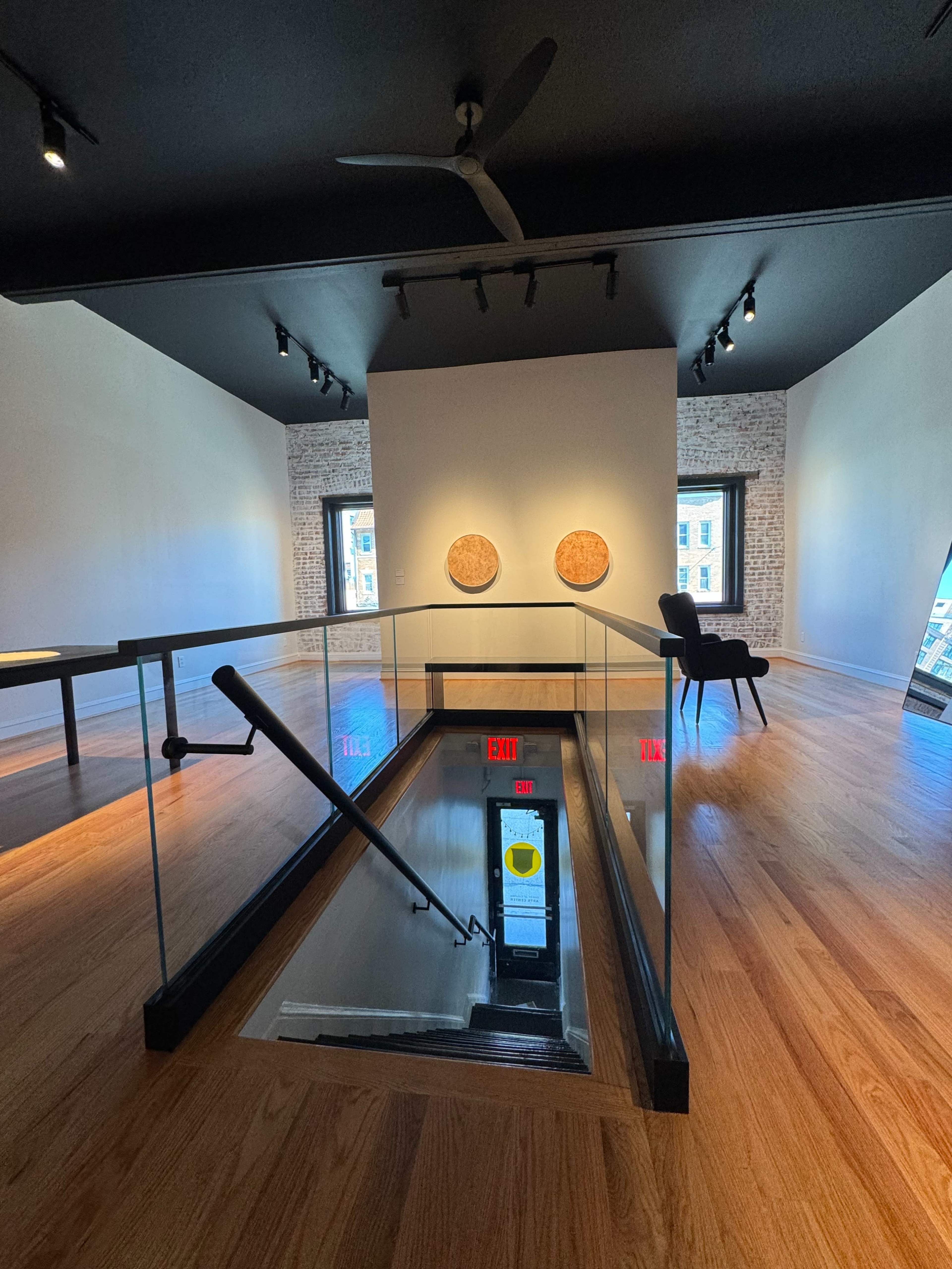 An interior art gallery featuring a glass railing staircase leading down to a lower level, with two circular artworks on the wall and a chair positioned nearby.