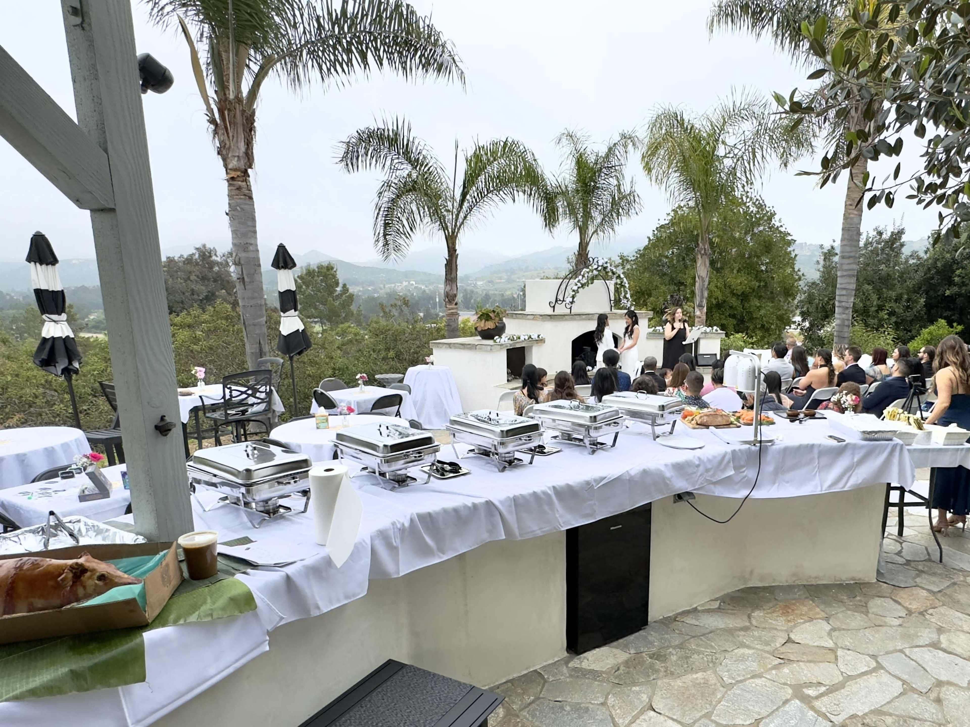 A buffet setup with trays of food is arranged on a table near a patio overlooking a scenic landscape, while guests gather for an event.
