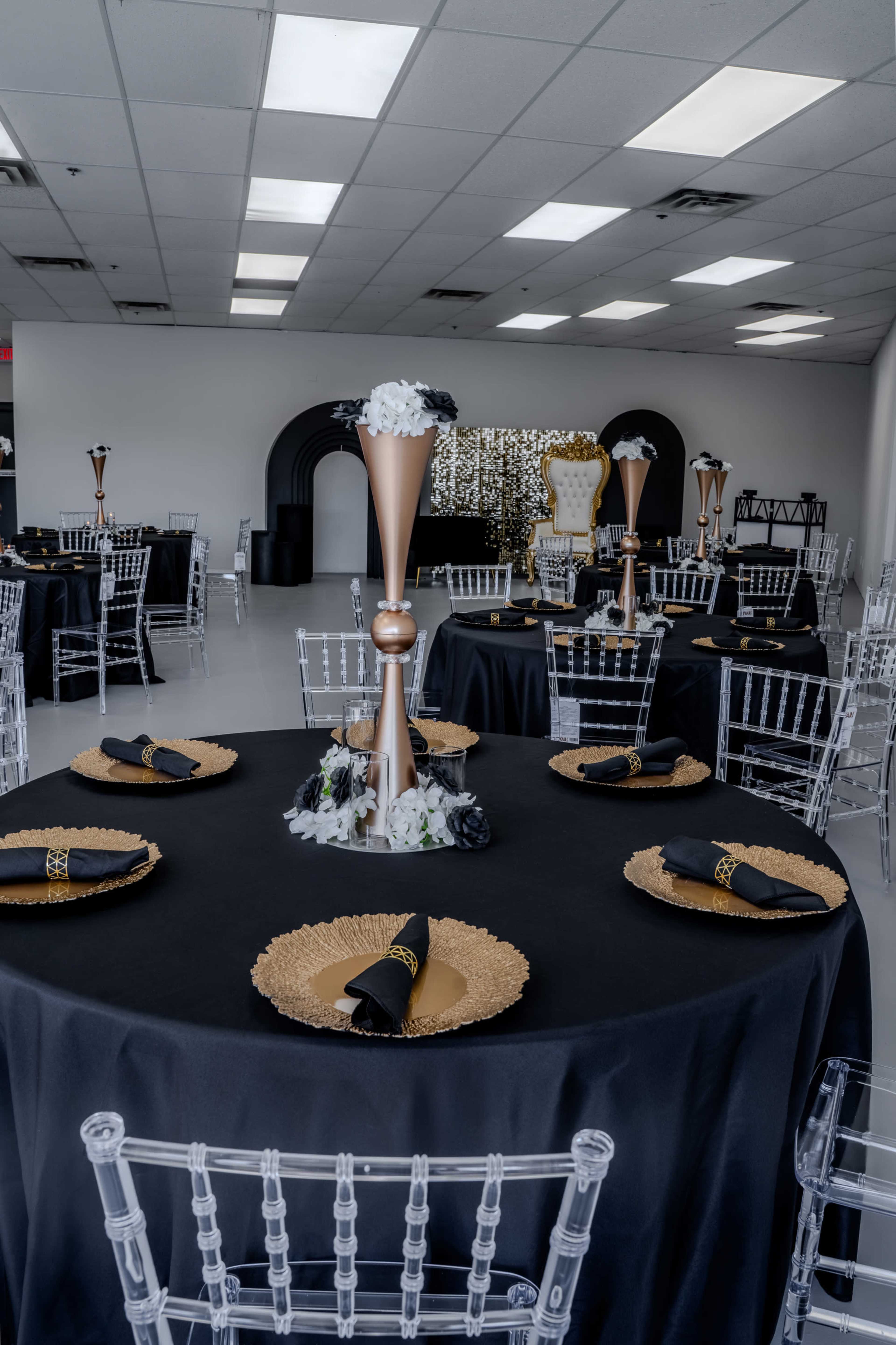 A decorated event space features black tables with gold charger plates, clear chairs, and elegant centerpieces.