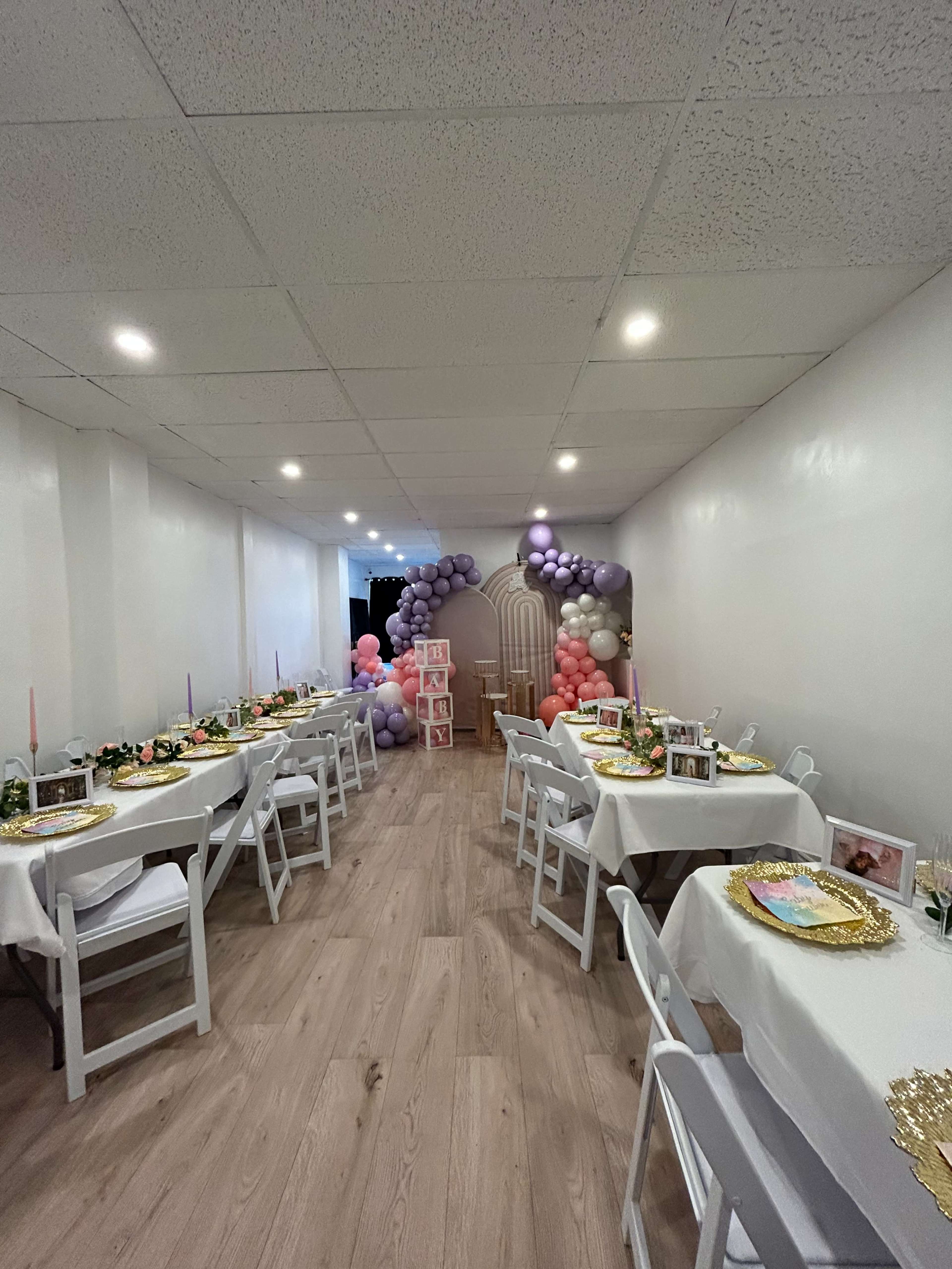 A spacious event room decorated with tables set for a celebration, featuring floral centerpieces and a balloon arch in the background.
