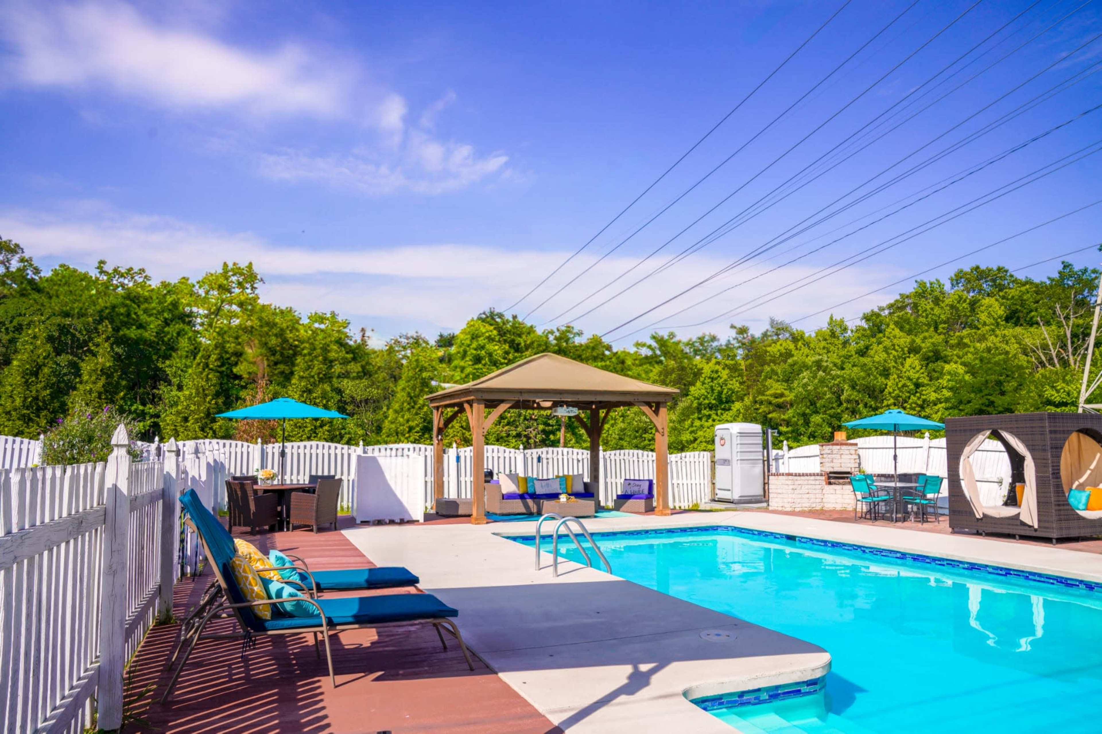 The image shows a swimming pool surrounded by a wooden deck, lounge chairs, a cabana, and green trees in the background.