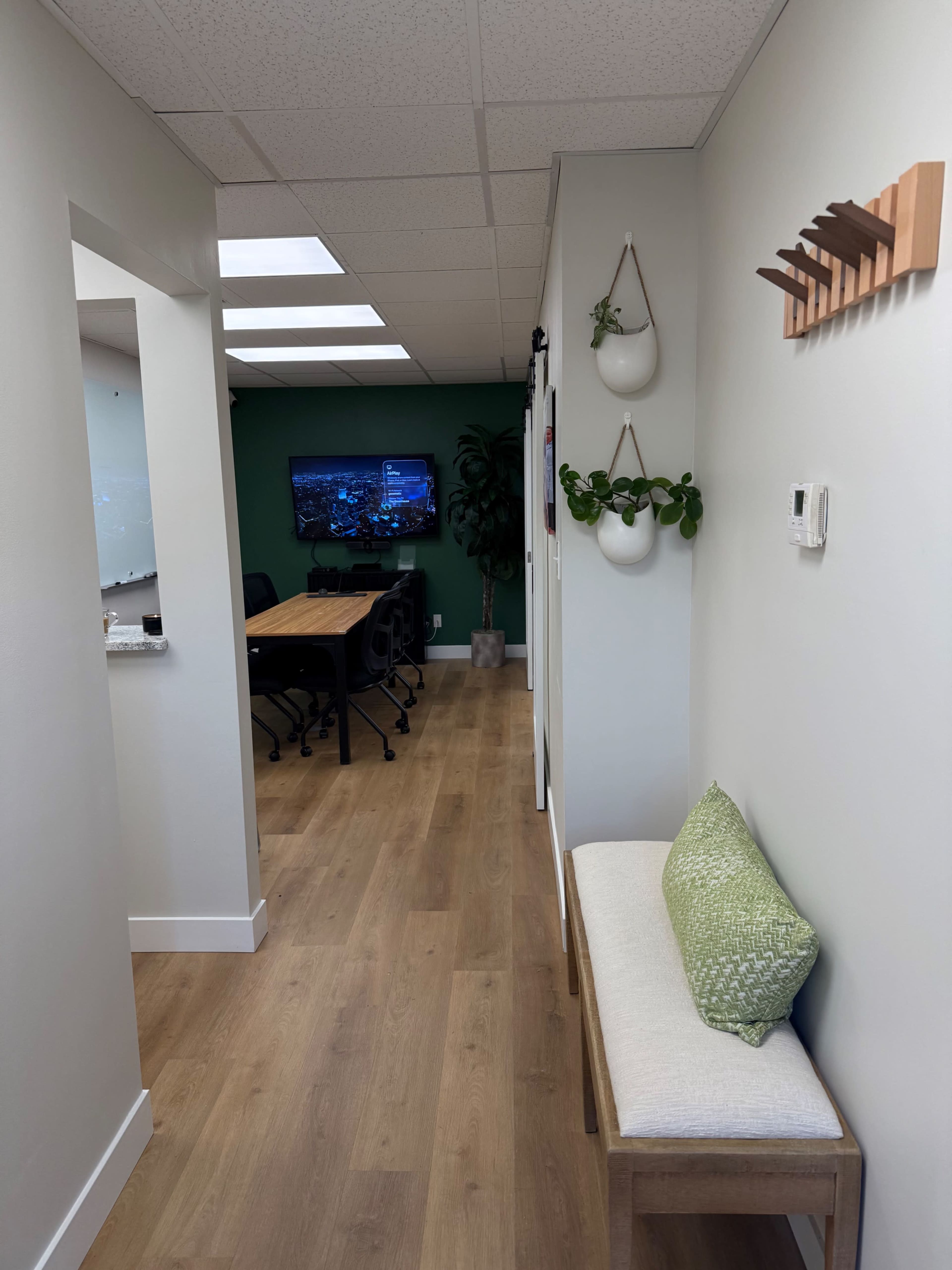 The image shows a hallway leading to a meeting room, with a bench on one side, wall plants, and a television displaying a cityscape in the background.
