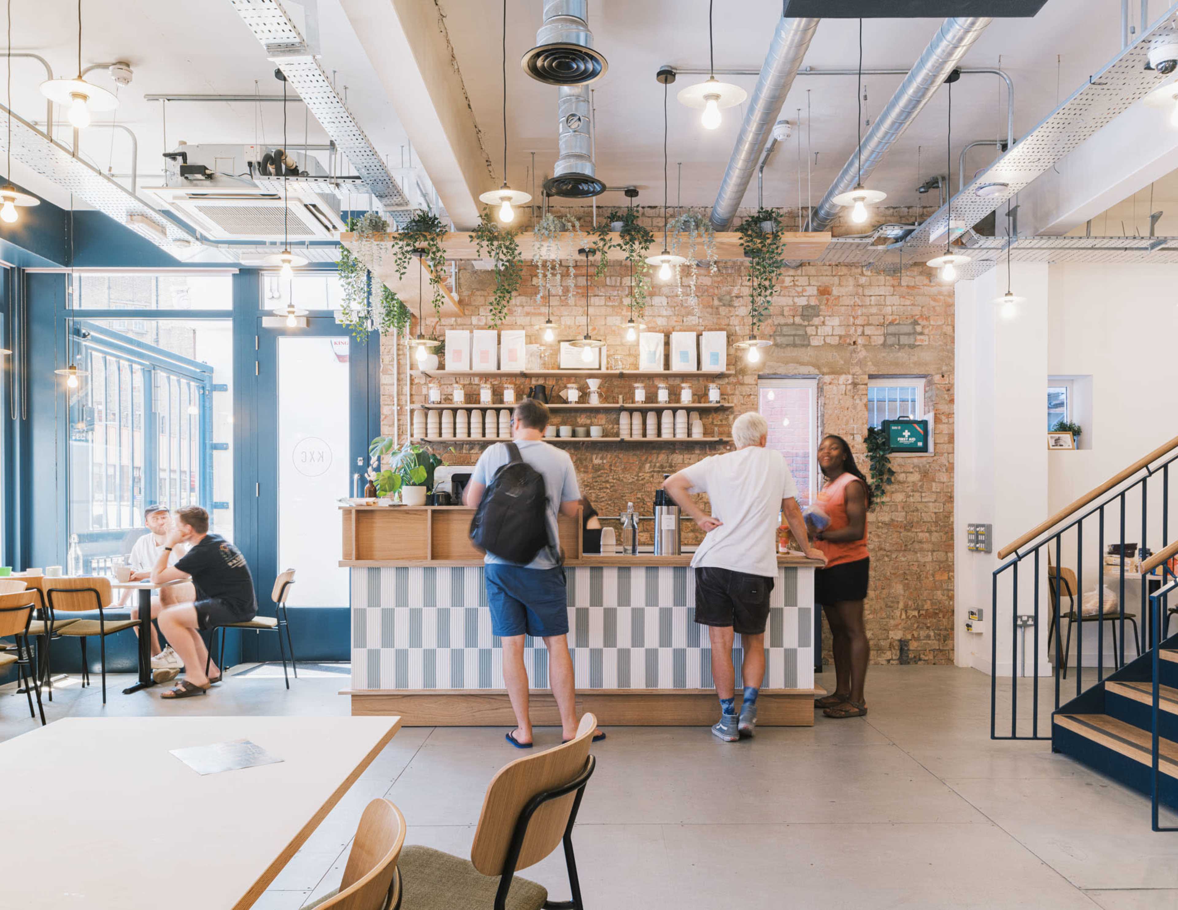 A café interior with a wooden counter, patrons ordering, and a mix of seating areas including tables and chairs, set against a backdrop of exposed brick walls and hanging light fixtures.