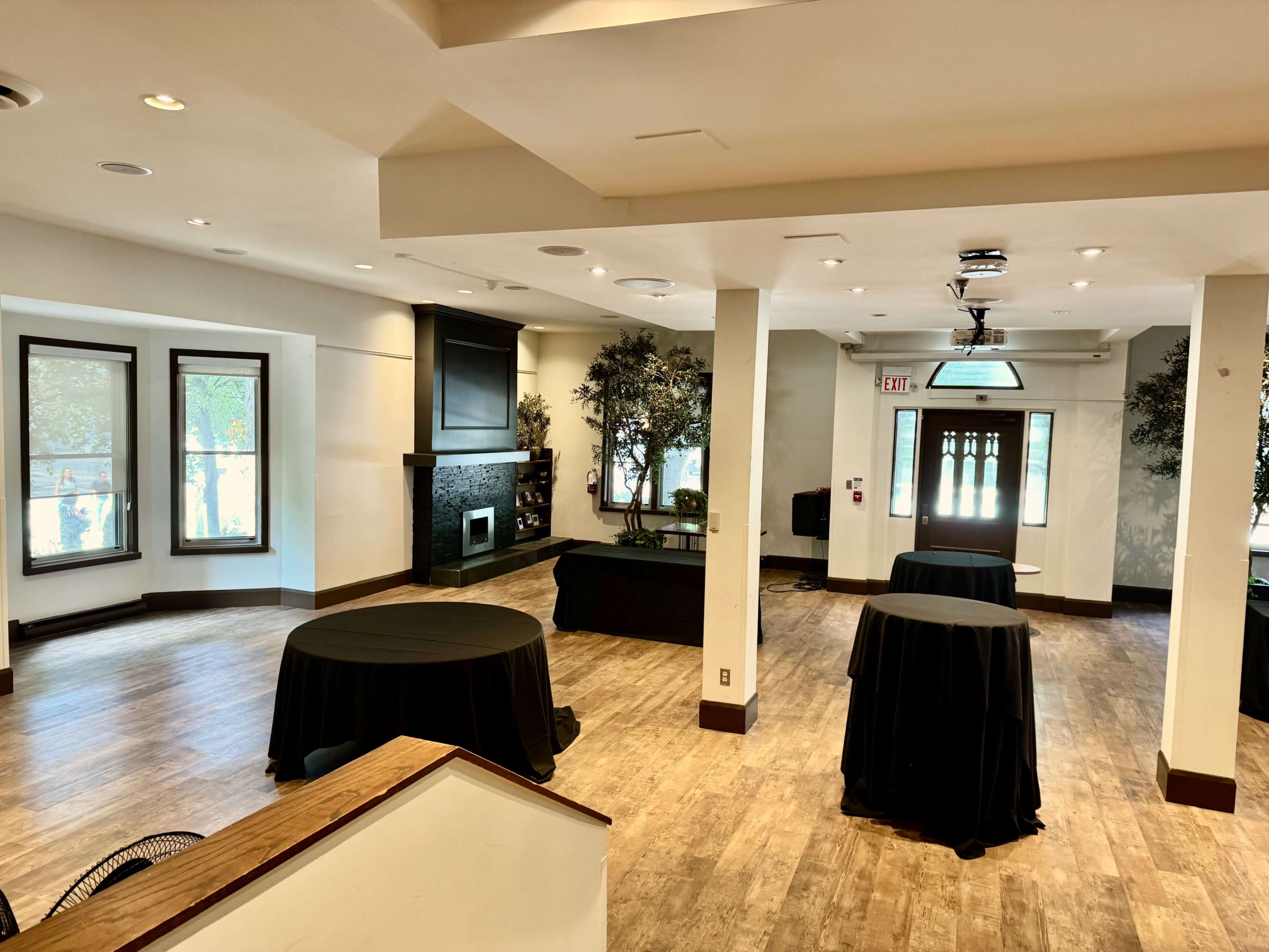 The image shows a spacious, well-lit room with wooden flooring, a fireplace on one wall, and several round tables covered with black tablecloths.
