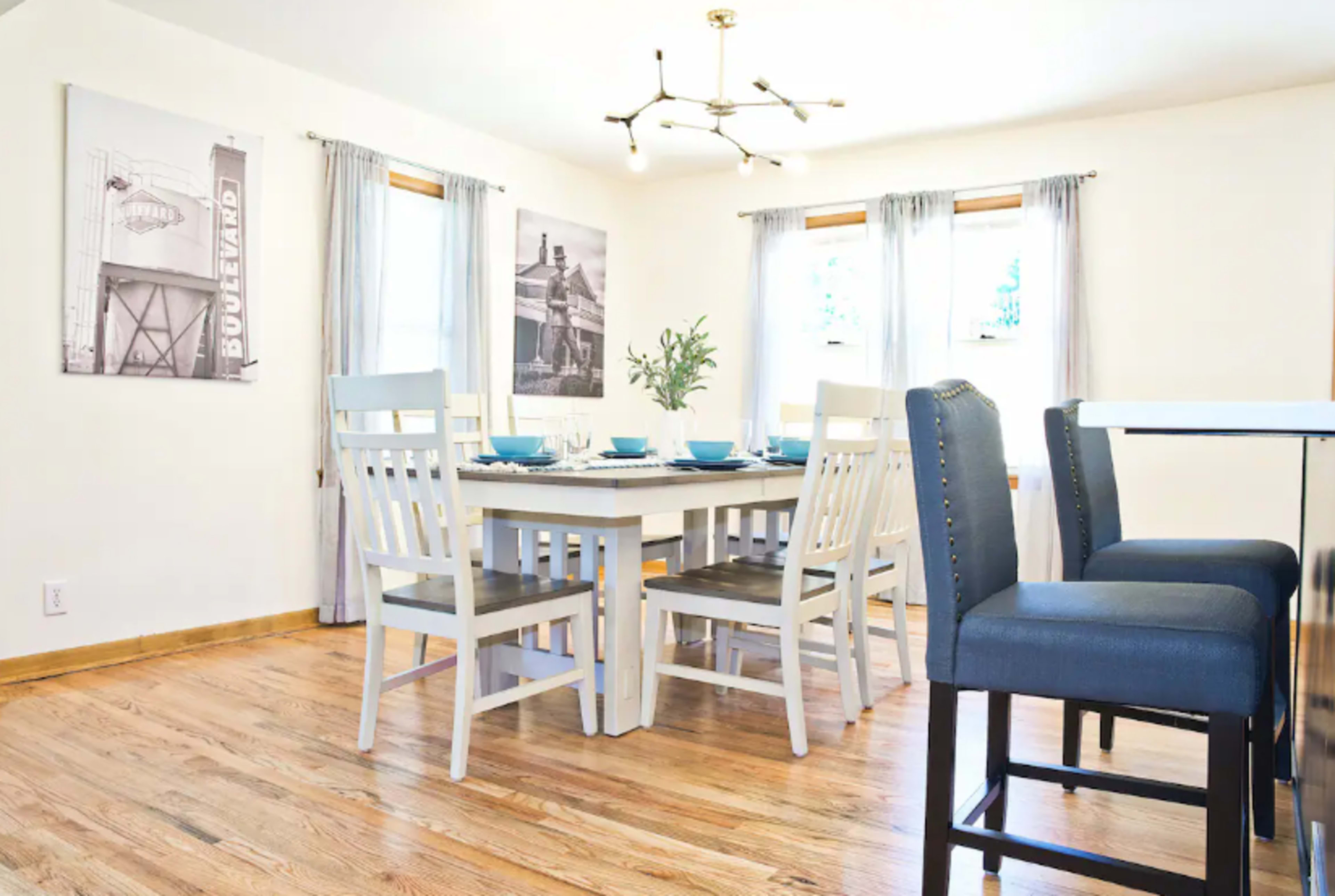 The image shows a modern dining area with a wooden table surrounded by six white chairs, complemented by a light fixture and large windows adorned with sheer curtains.