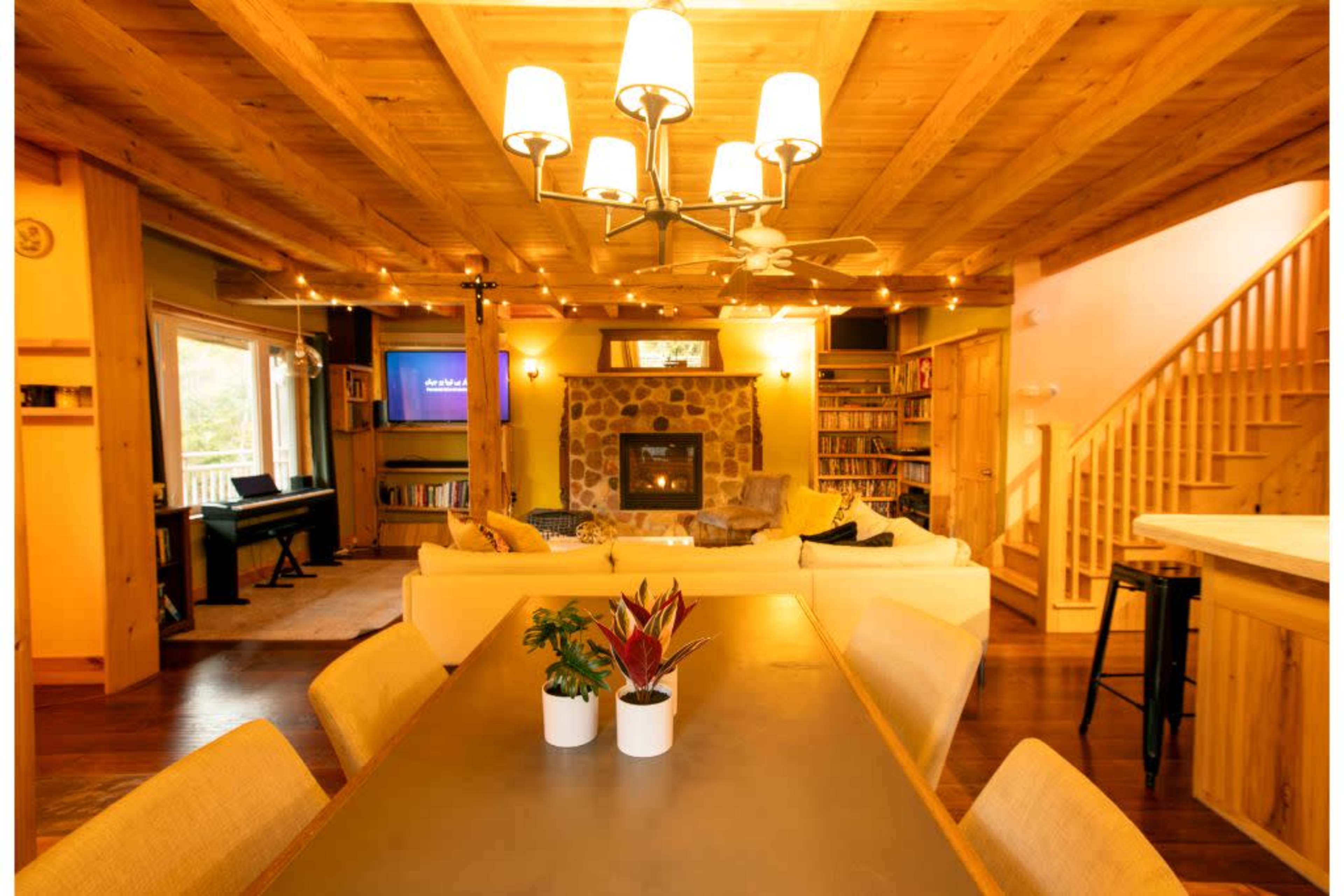 A spacious living area features a stone fireplace, a large sectional sofa, a dining table with potted plants, and wooden beams on the ceiling.