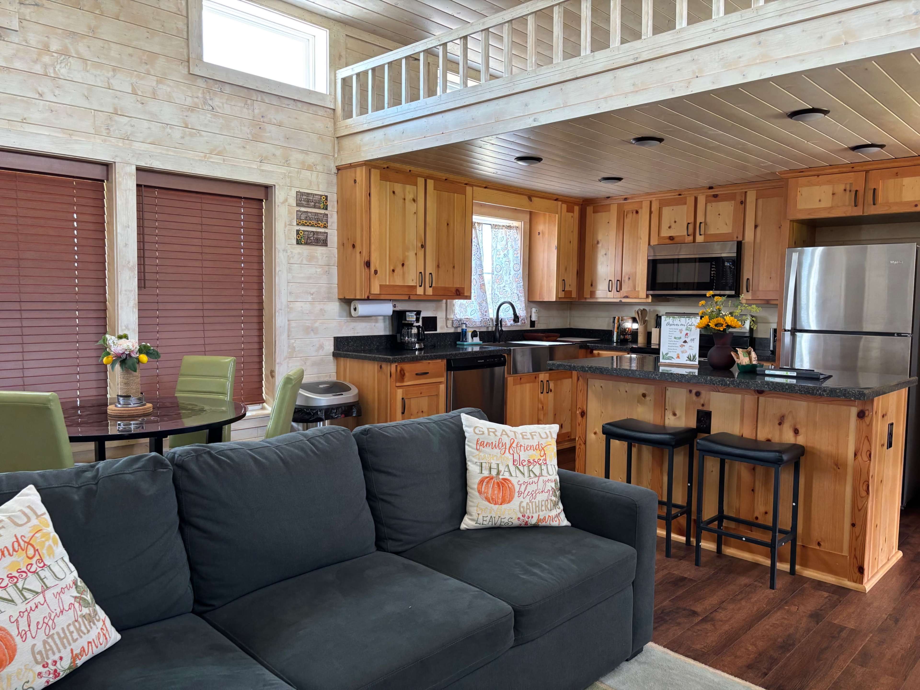 The image shows a cozy interior of a wooden cabin with a kitchen and living area featuring a dark couch and a dining table.