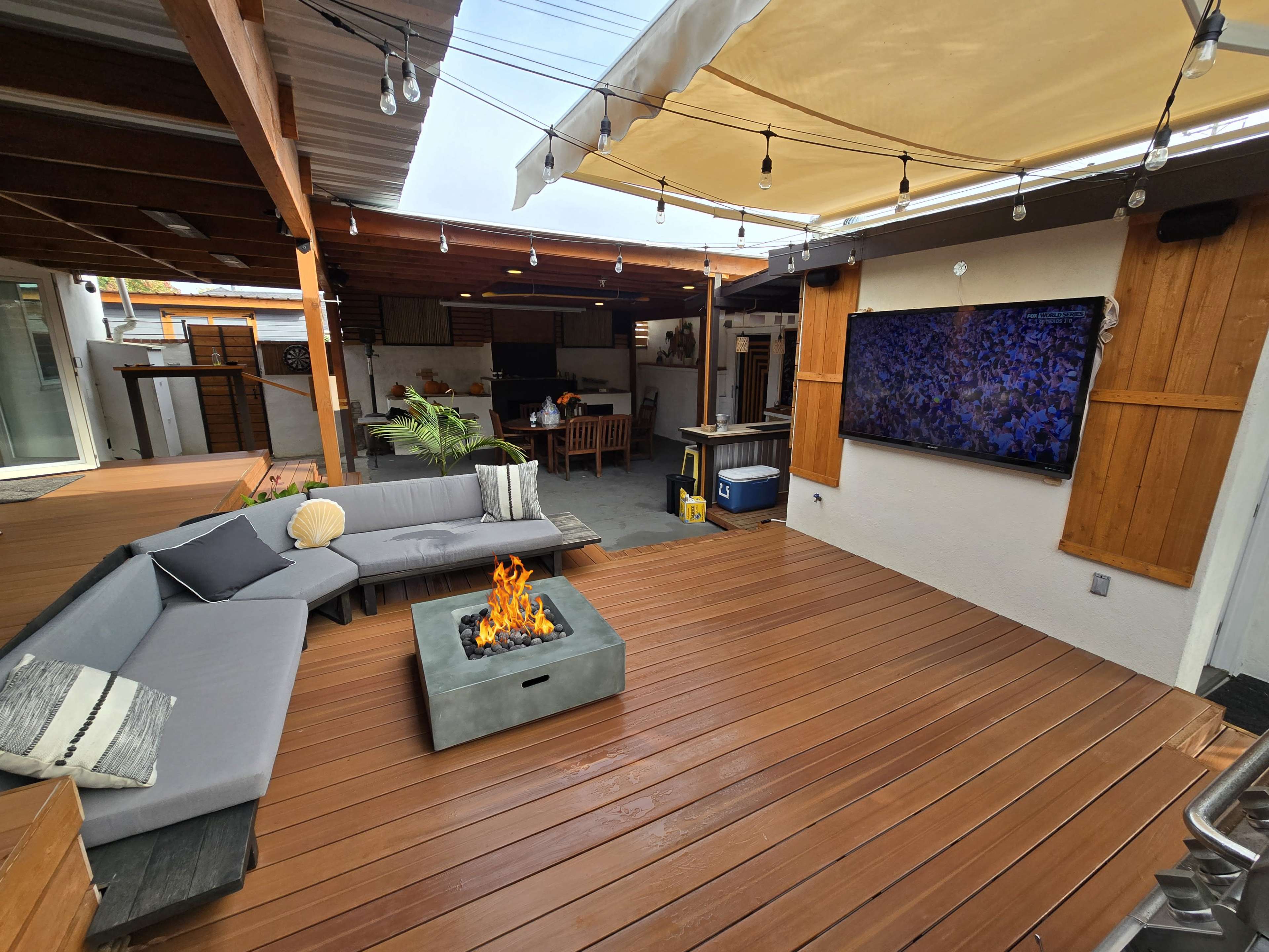 The outdoor patio features a modern sectional sofa, a fire pit in the center, an outdoor kitchen area, and a mounted television.