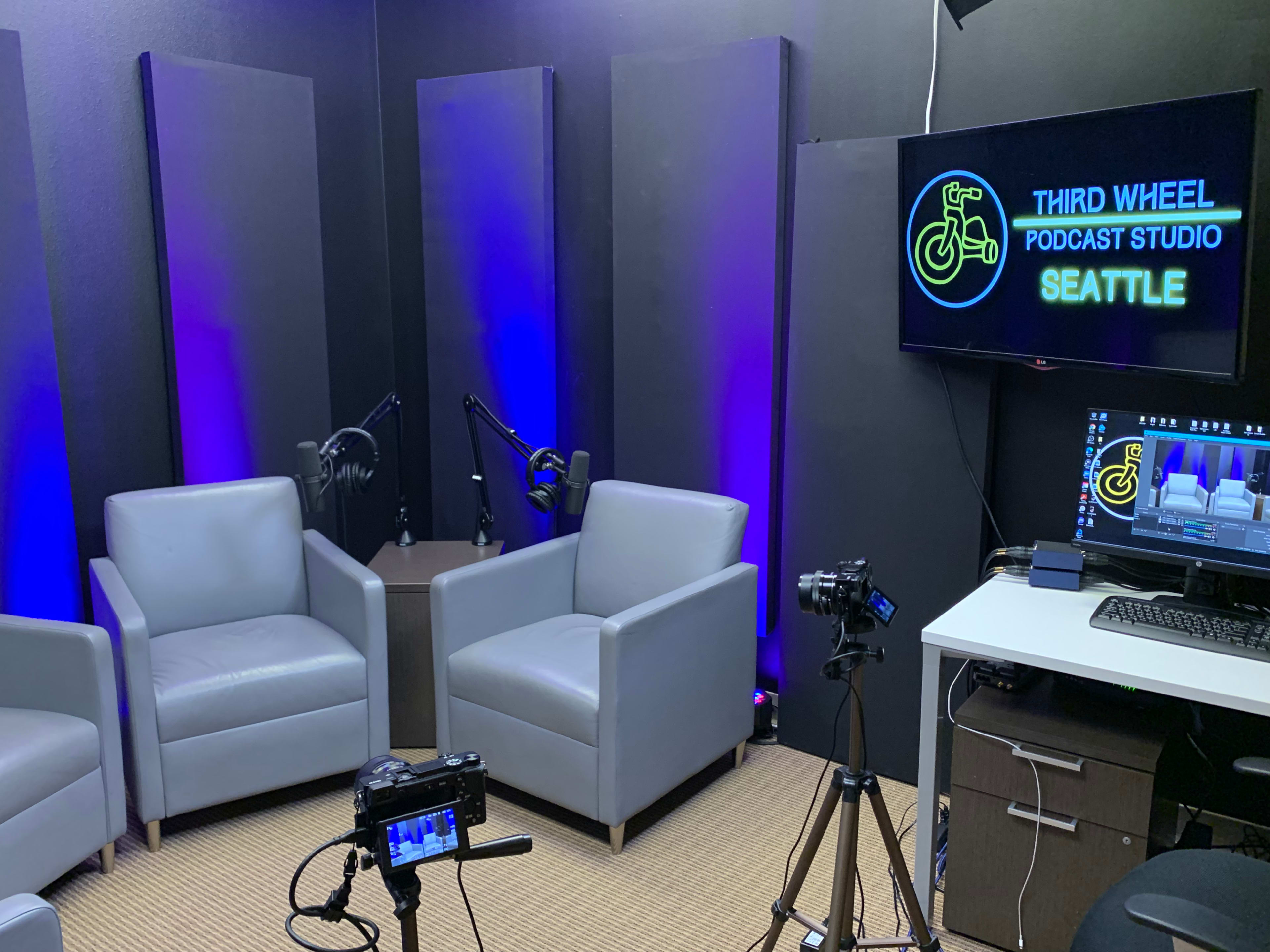 A podcast studio featuring two grey armchairs, a television displaying "Third Wheel Podcast Studio Seattle," and camera equipment set up on a table.