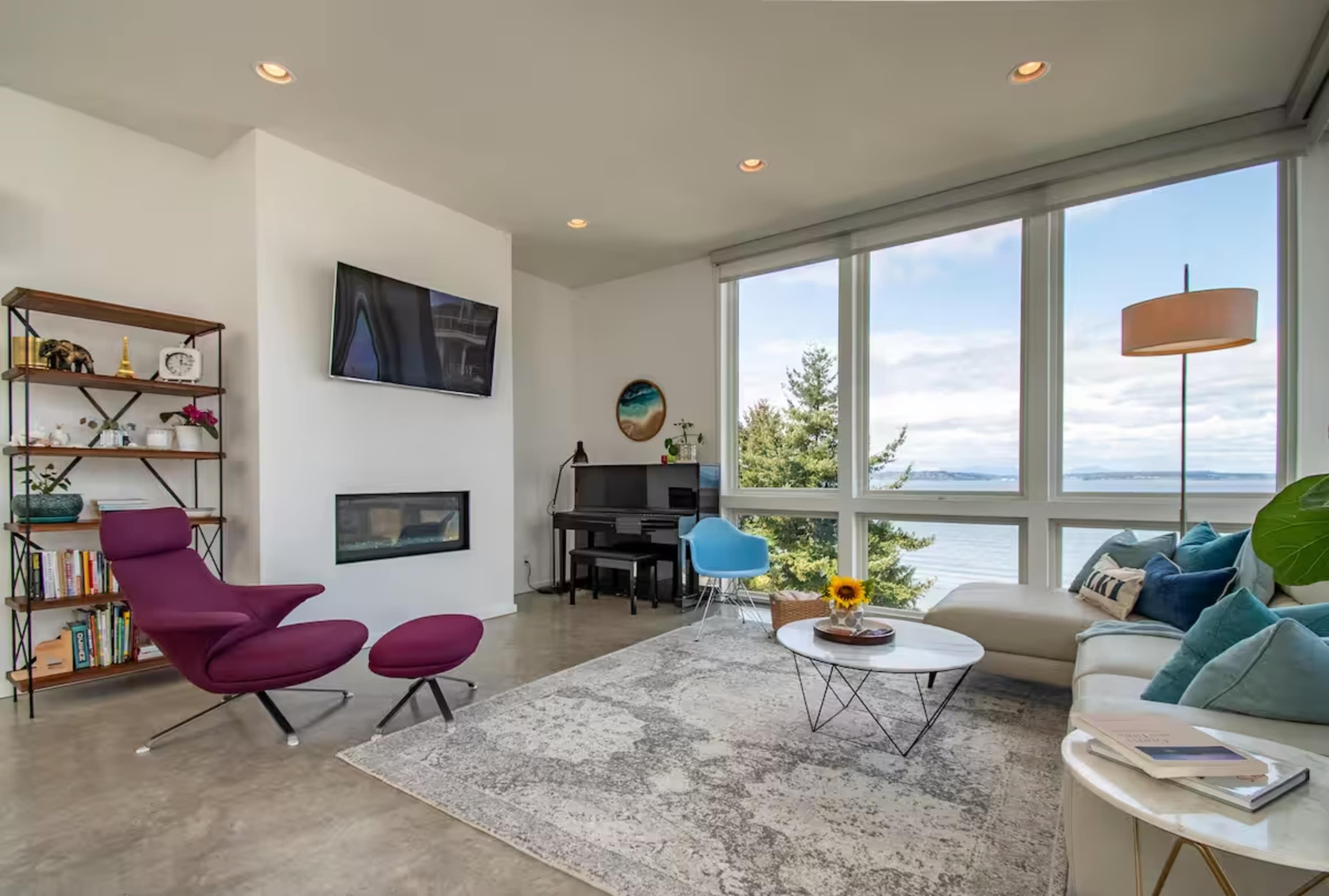 The image shows a modern living room featuring a purple chair, a white coffee table with a sunflower, a sofa with decorative pillows, a piano, and large windows offering a view of the outdoors.