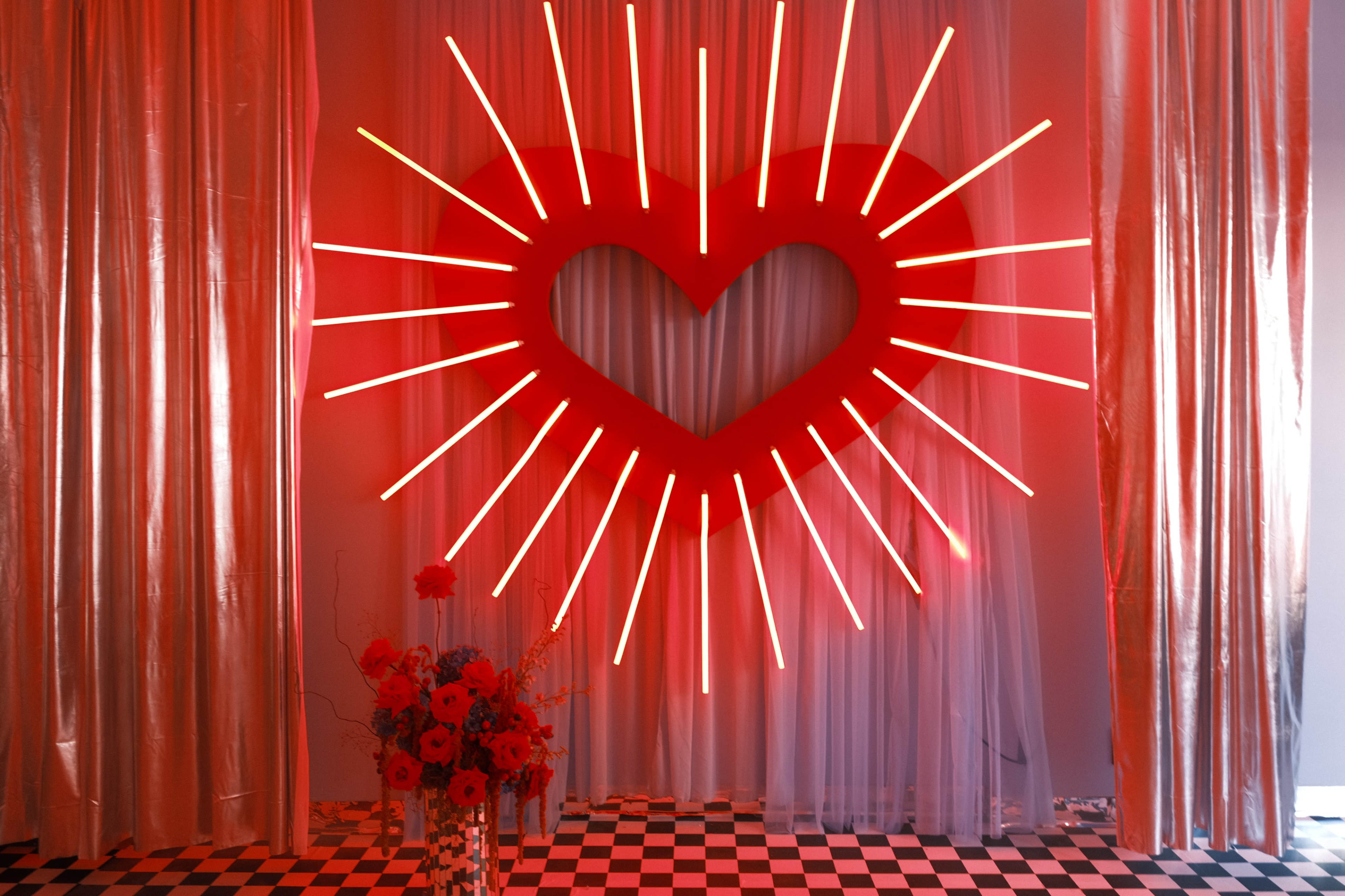 A large red heart-shaped neon light with rays radiating from it is displayed against a backdrop of red curtains and a black-and-white checkered floor, accompanied by a floral arrangement in shades of red.