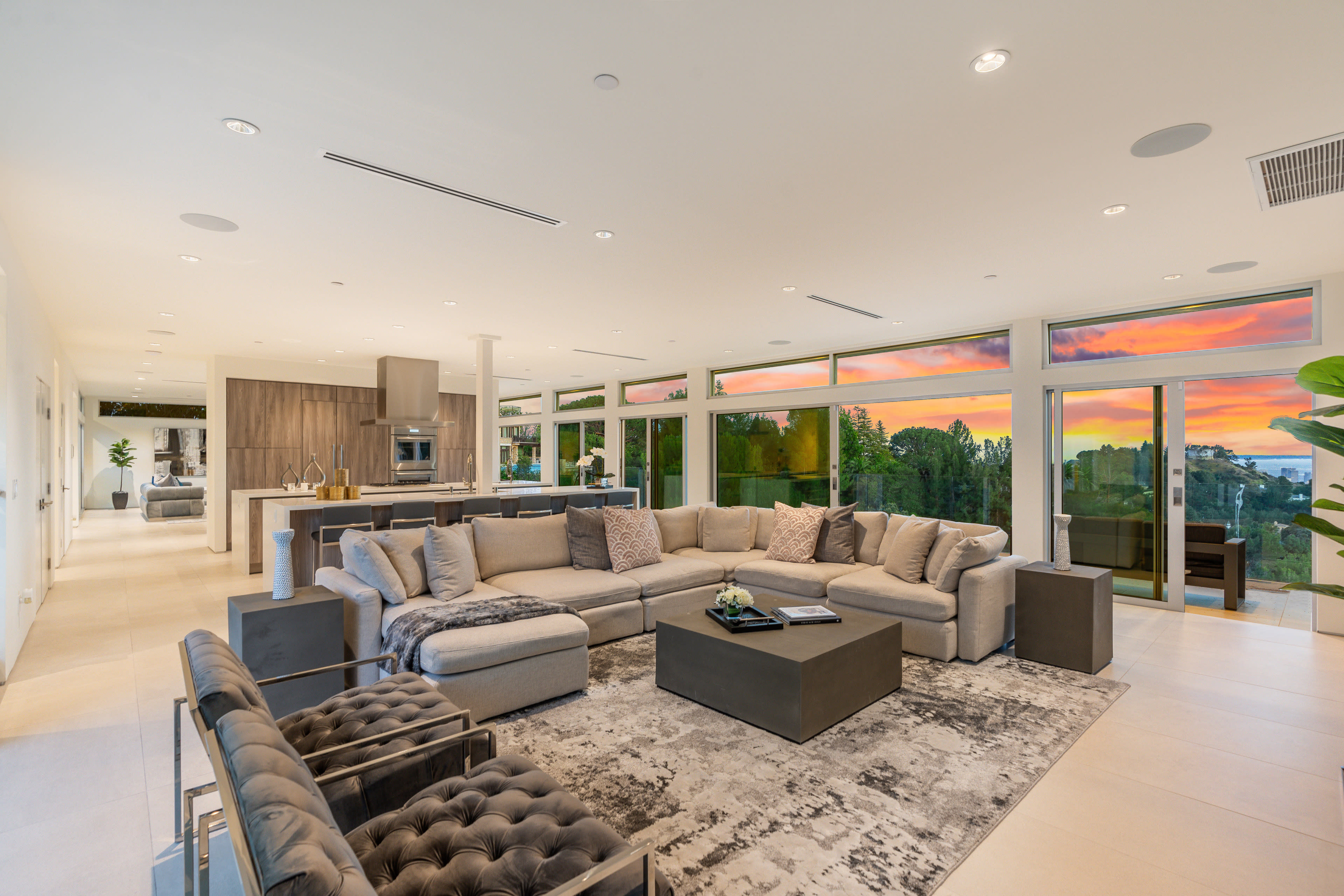 The image shows a spacious, modern living room with large windows overlooking a colorful sunset, complemented by contemporary furniture and a cozy seating arrangement.