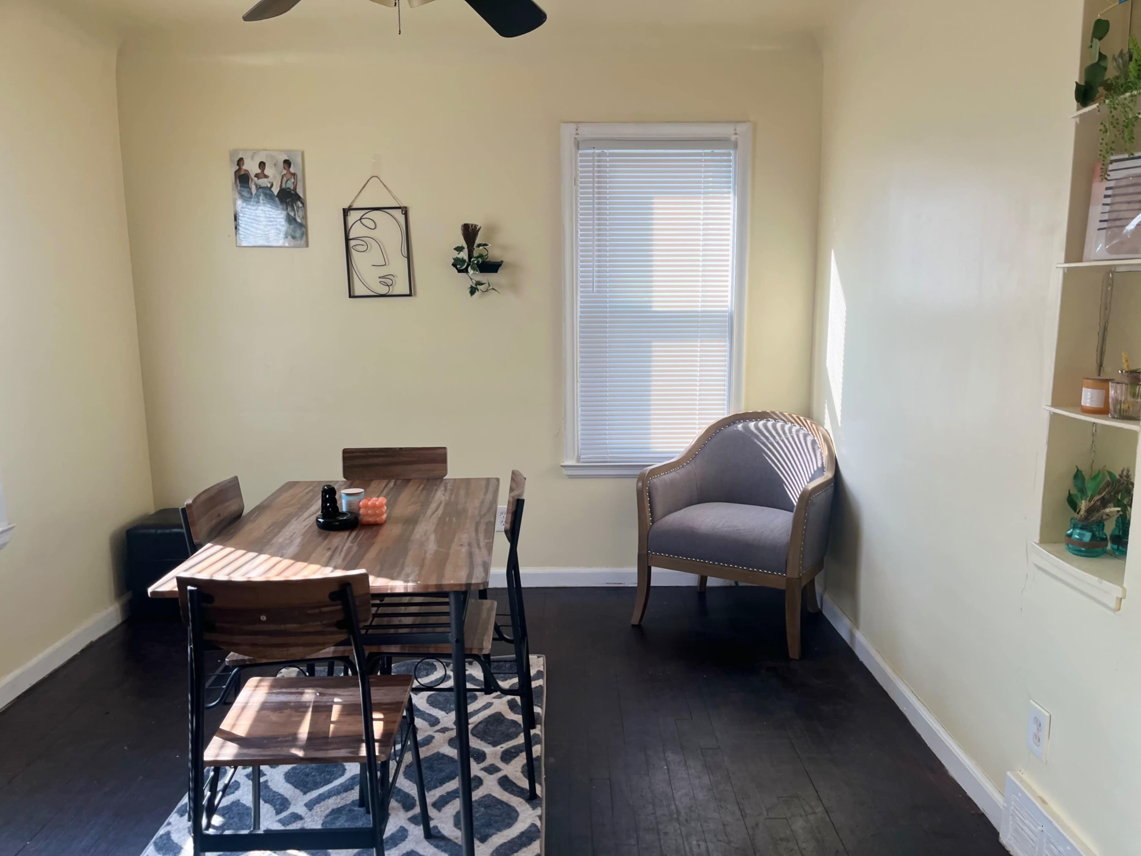 The image shows a small dining area with a wooden table, four chairs, a window with blinds, and a single armchair in a room with light yellow walls.