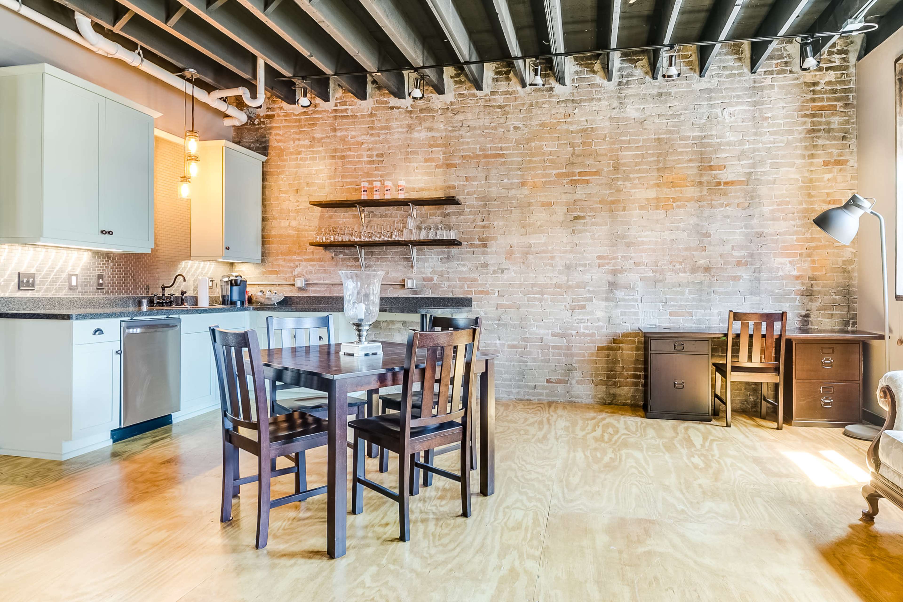 The image shows a modern kitchen and dining area with exposed brick walls, a wooden dining table surrounded by chairs, and a small desk in the corner.